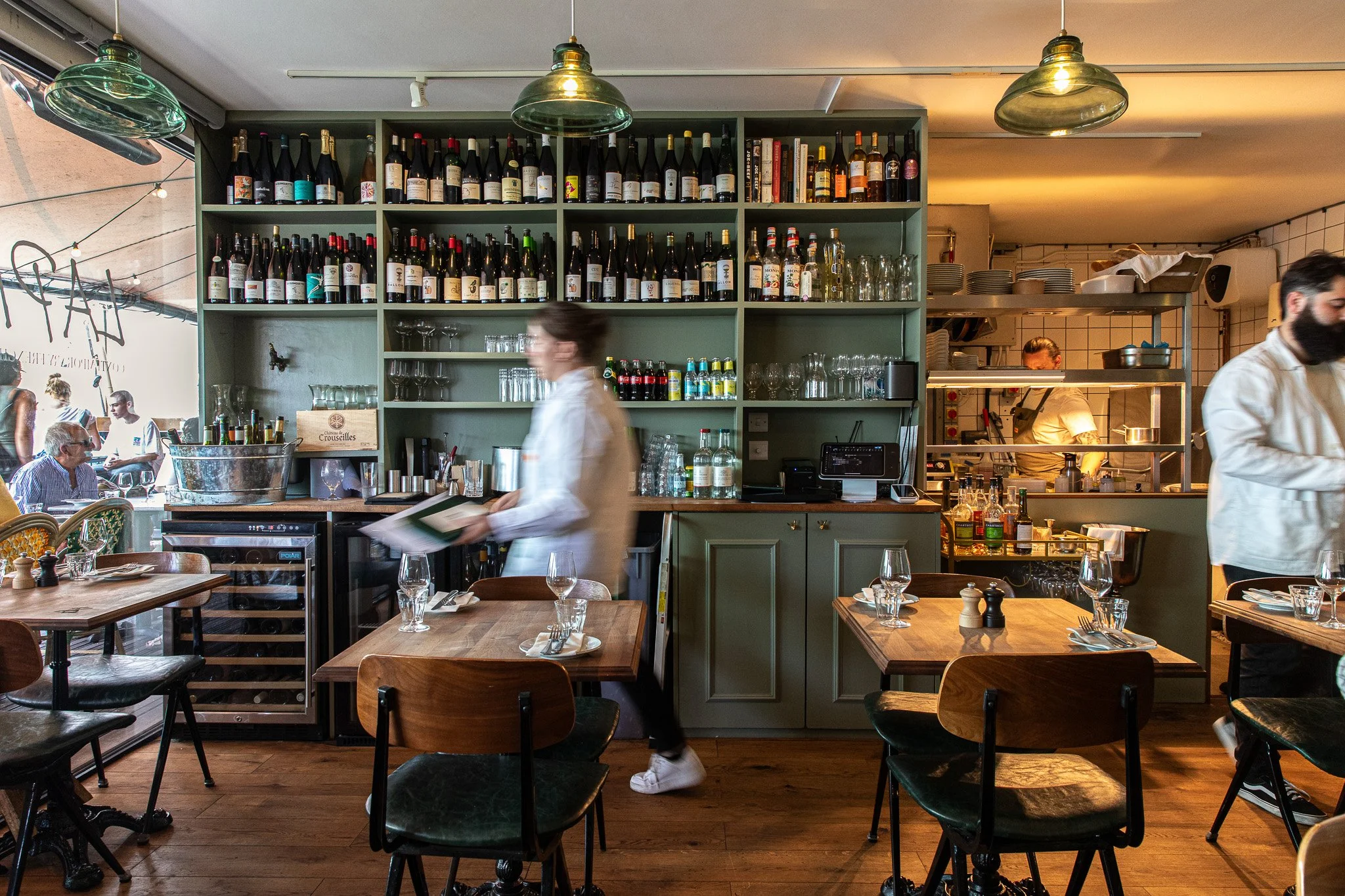 Interior of a restaurant with a green wine shelf behind the bar, wooden tables set with plates, glasses, and utensils, staff wearing white shirts working, and chefs in the kitchen visible through an open window.