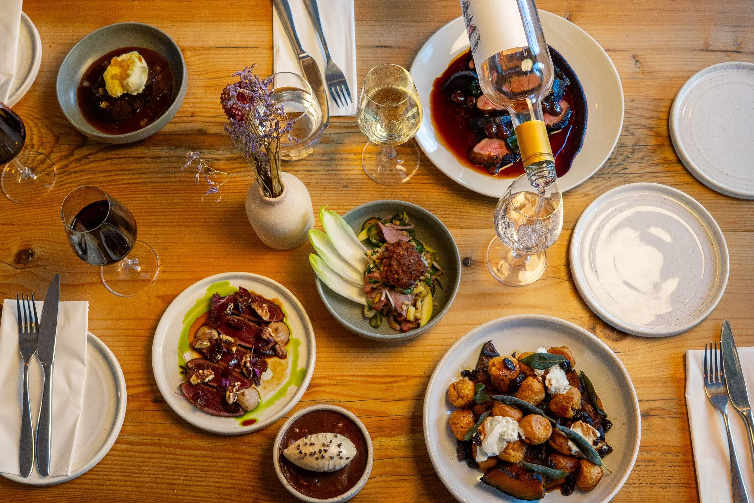 A wooden dining table set with various dishes including a salad, roasted vegetables, meat, and dessert, along with wine glasses and a small vase with purple flowers.