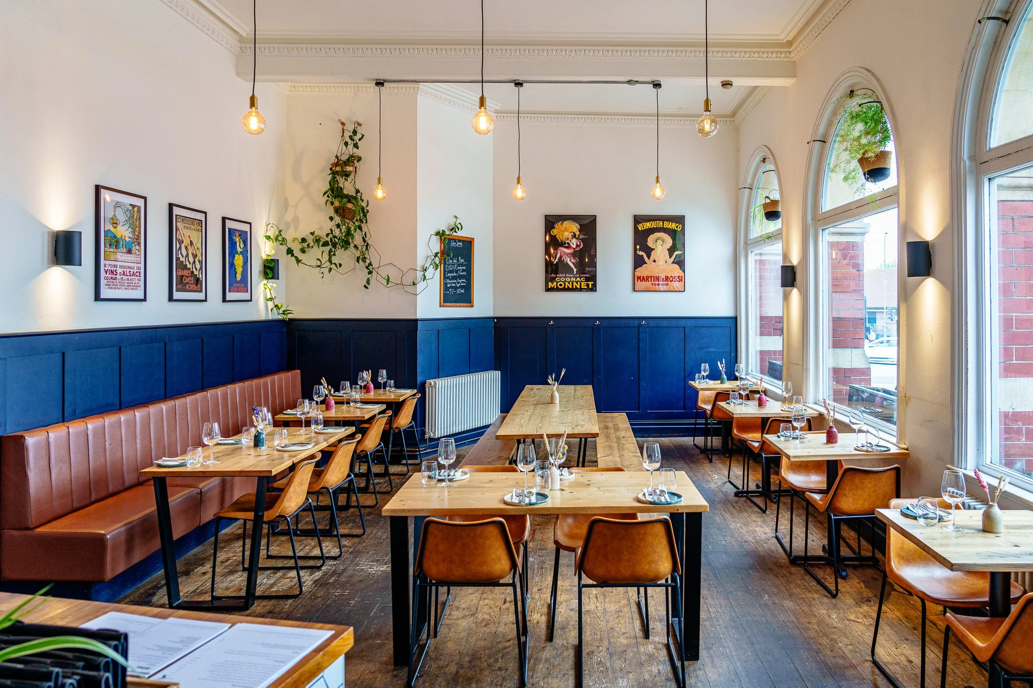Interior of a cozy, well-lit restaurant with wooden tables and chairs, large windows, framed vintage posters on the walls, hanging light bulbs, and plants.