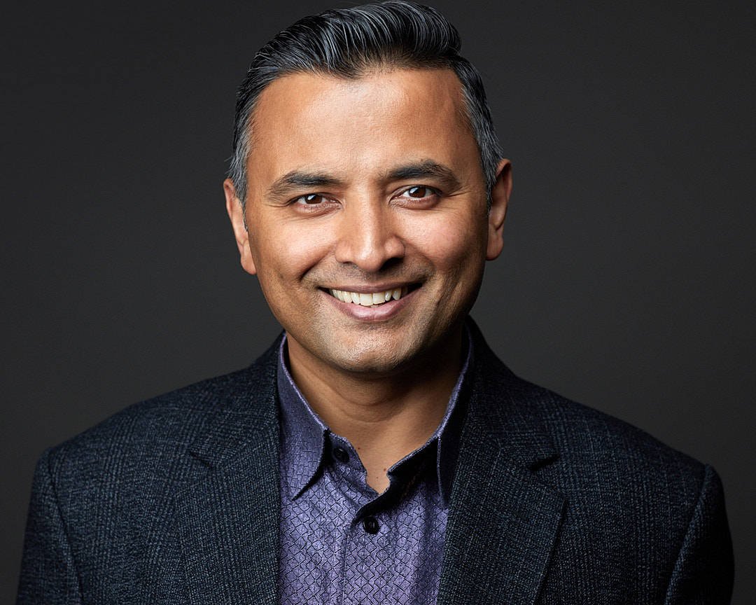 Clarksburg headshot of a smiling man in dark suit and patterned shirt against dark background.