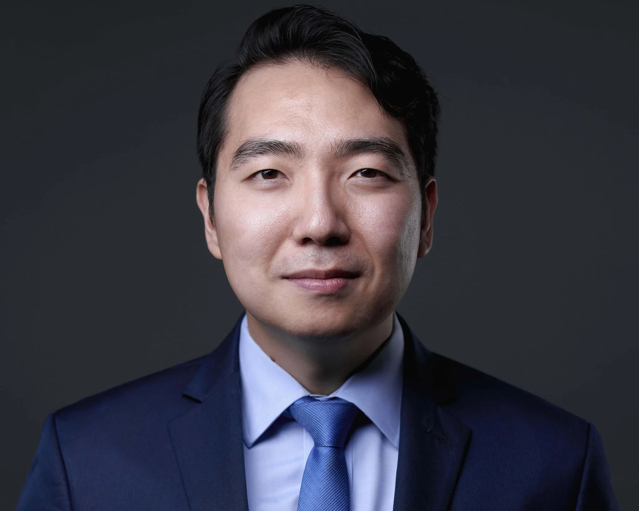 Professional headshot of a young Asian man with dark hair, wearing a navy suit, light blue shirt, and matching blue tie, against a dark gray background.
