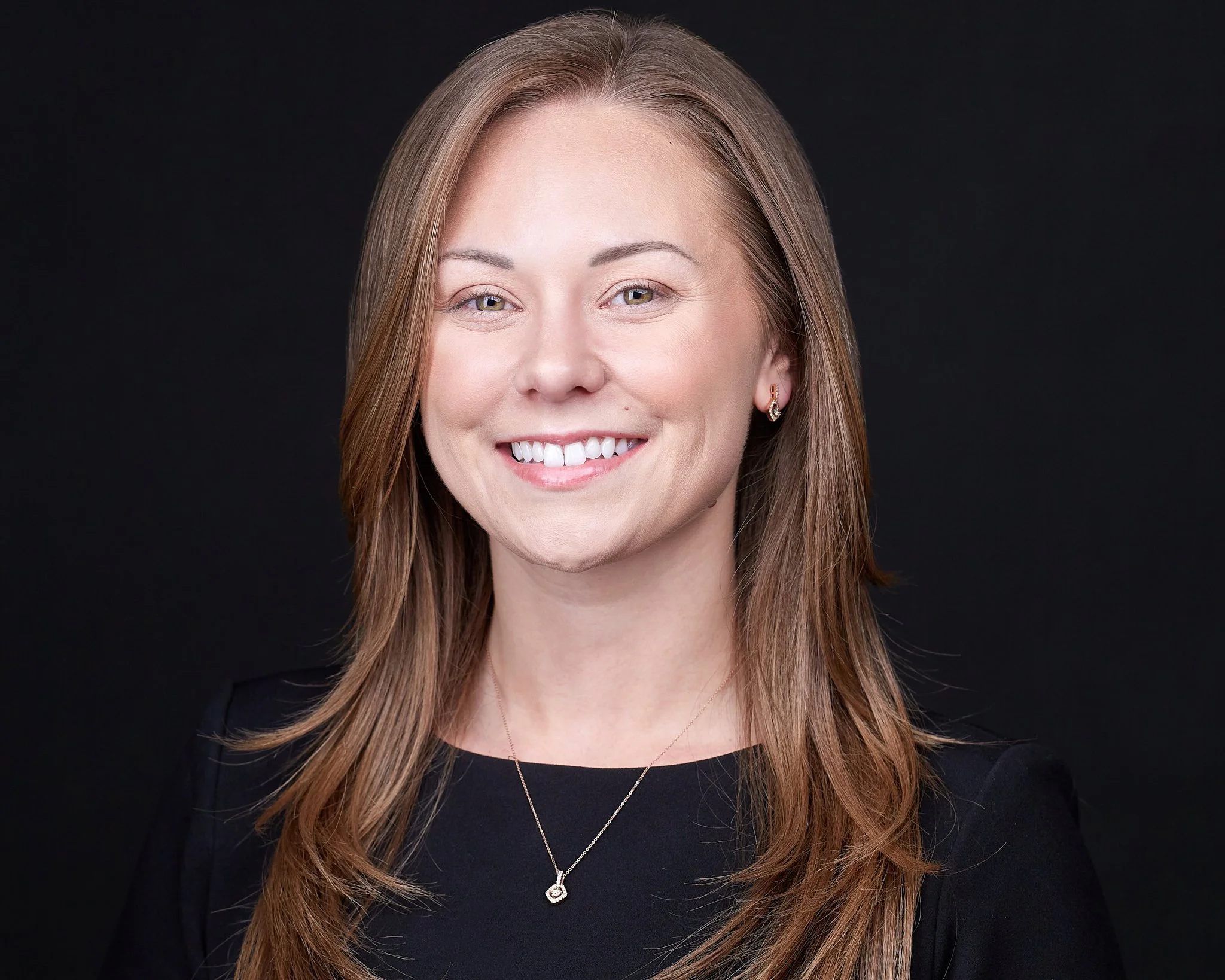 Headshot of a smiling woman with light skin and long, straight, light brown hair, wearing a black top and a delicate necklace, against a dark background.
