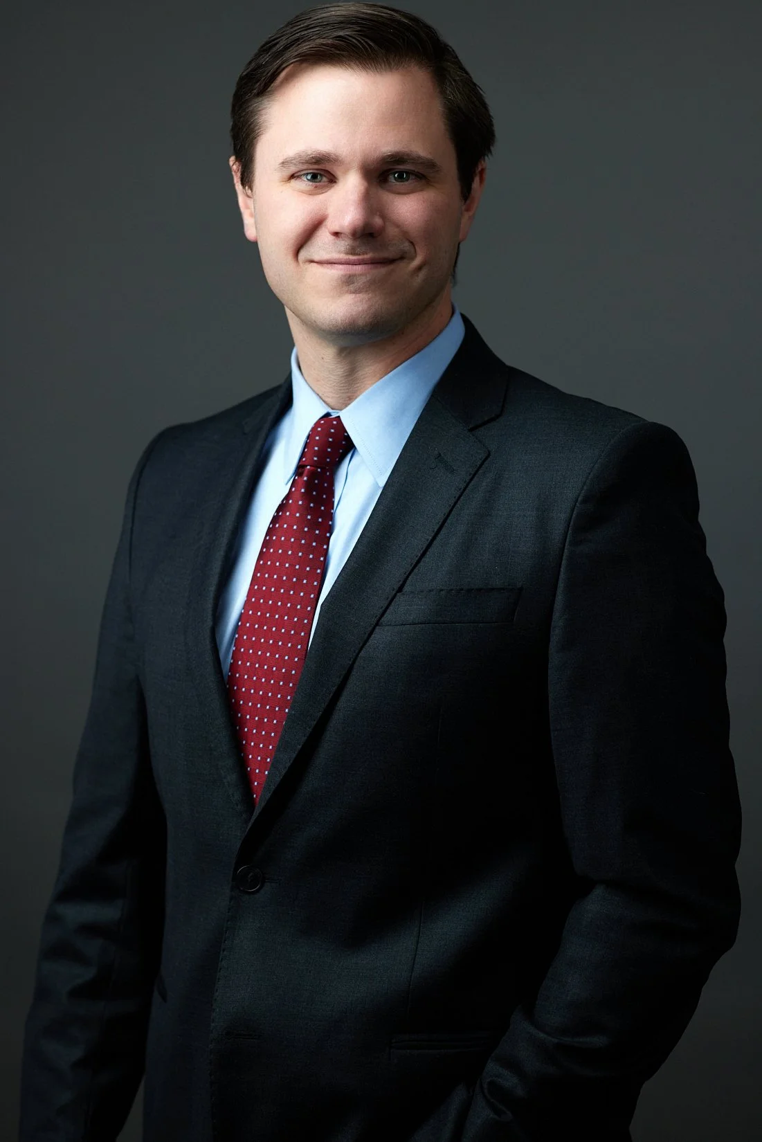Professional portrait of a man in a dark suit, light blue shirt, and red tie with white dots, smiling and standing against a dark gray background.