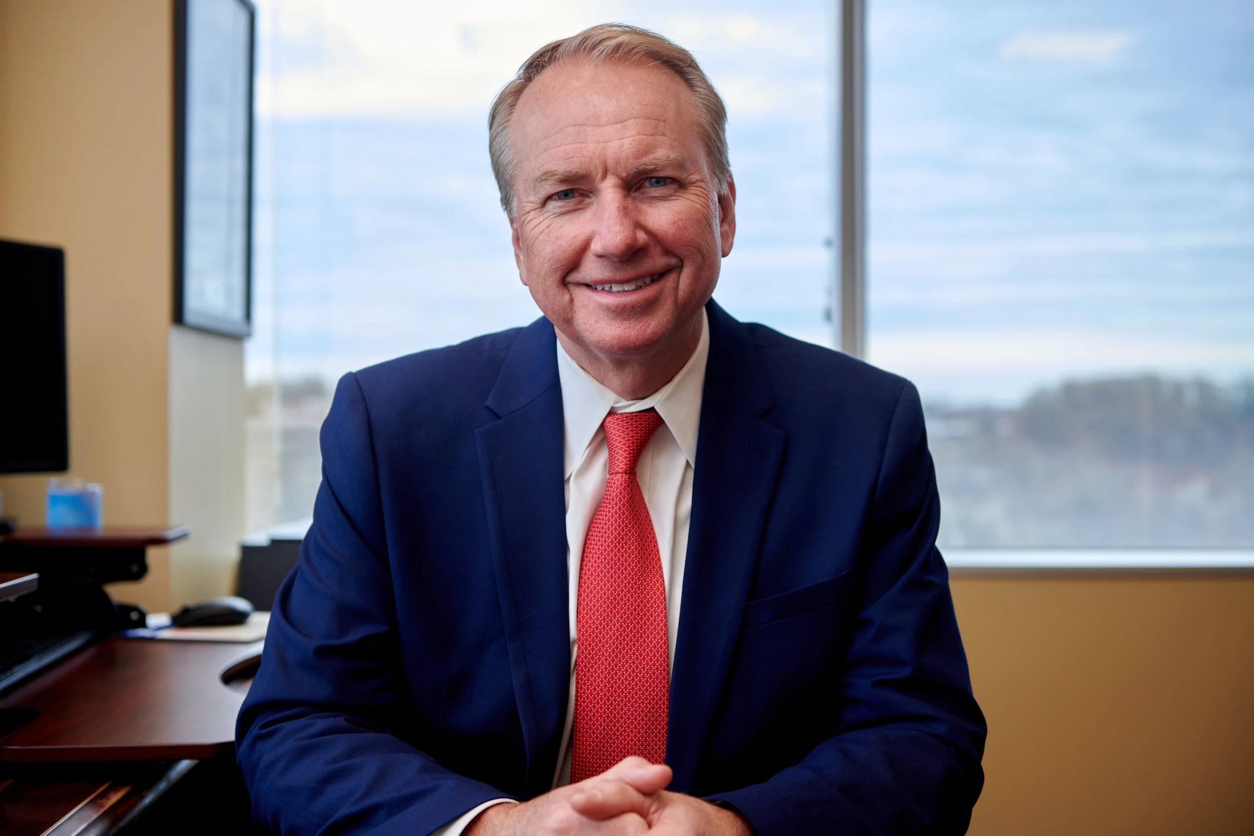 A smiling middle-aged man in a navy suit and red tie sitting at a desk in an office.