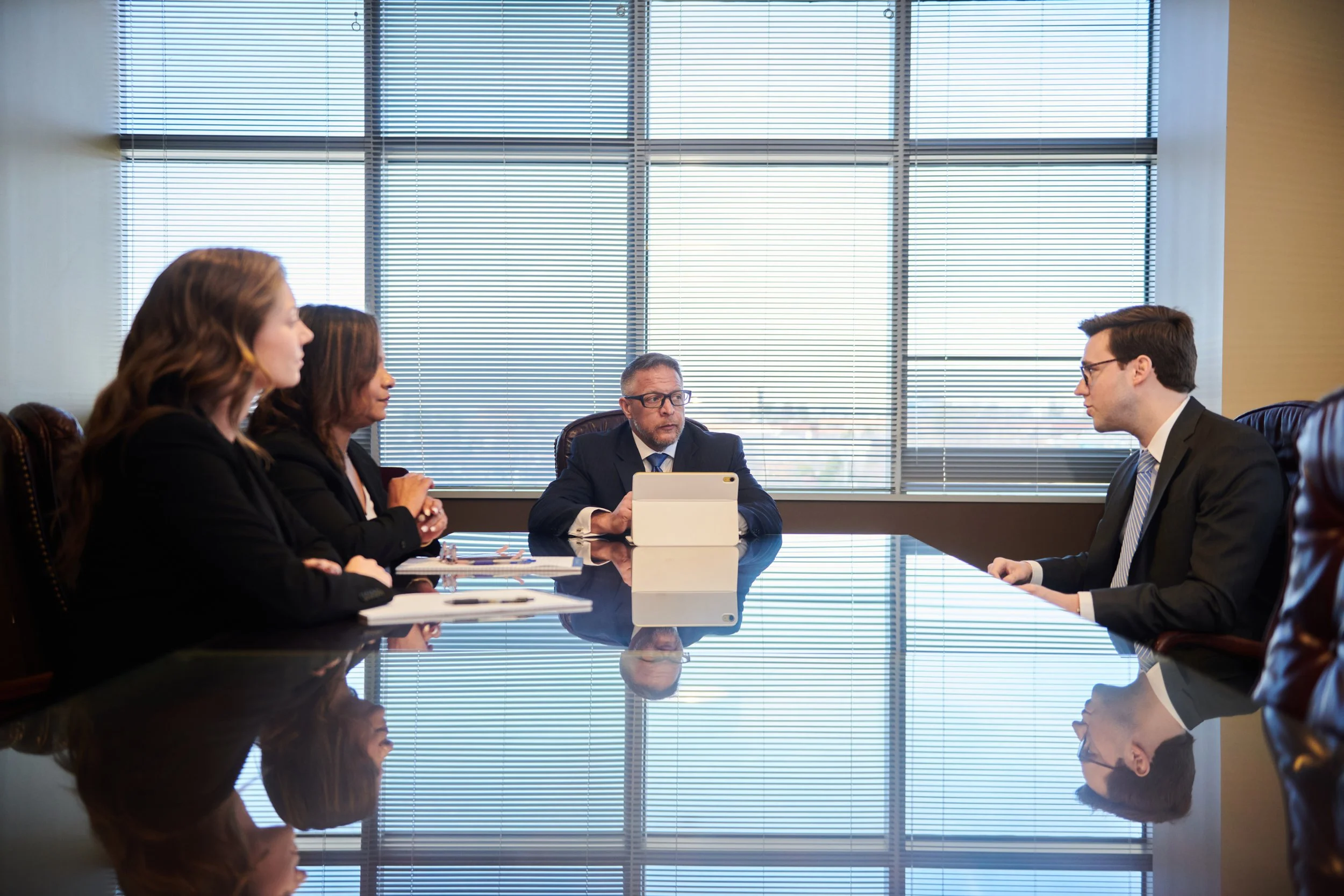 Business meeting in a modern office, with four people in suits sitting across a reflective conference table, engaged in discussion.