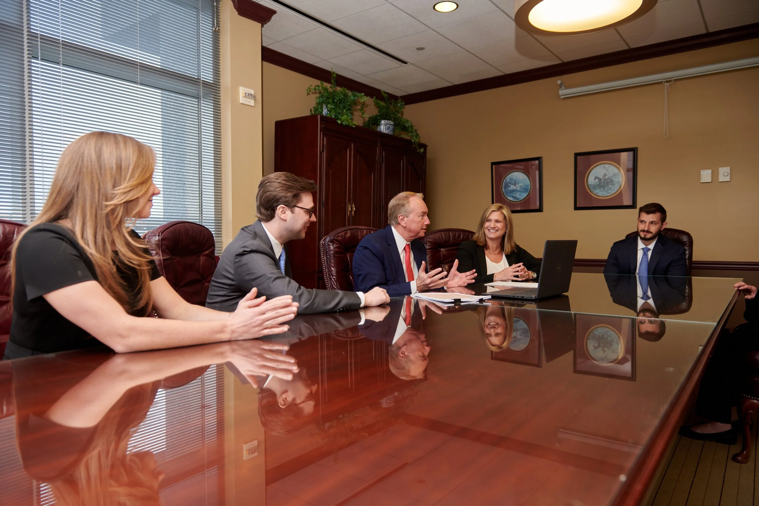 Business meeting with five professionals seated at a large, polished conference table in a room with beige walls, framed artwork, and a window with blinds.