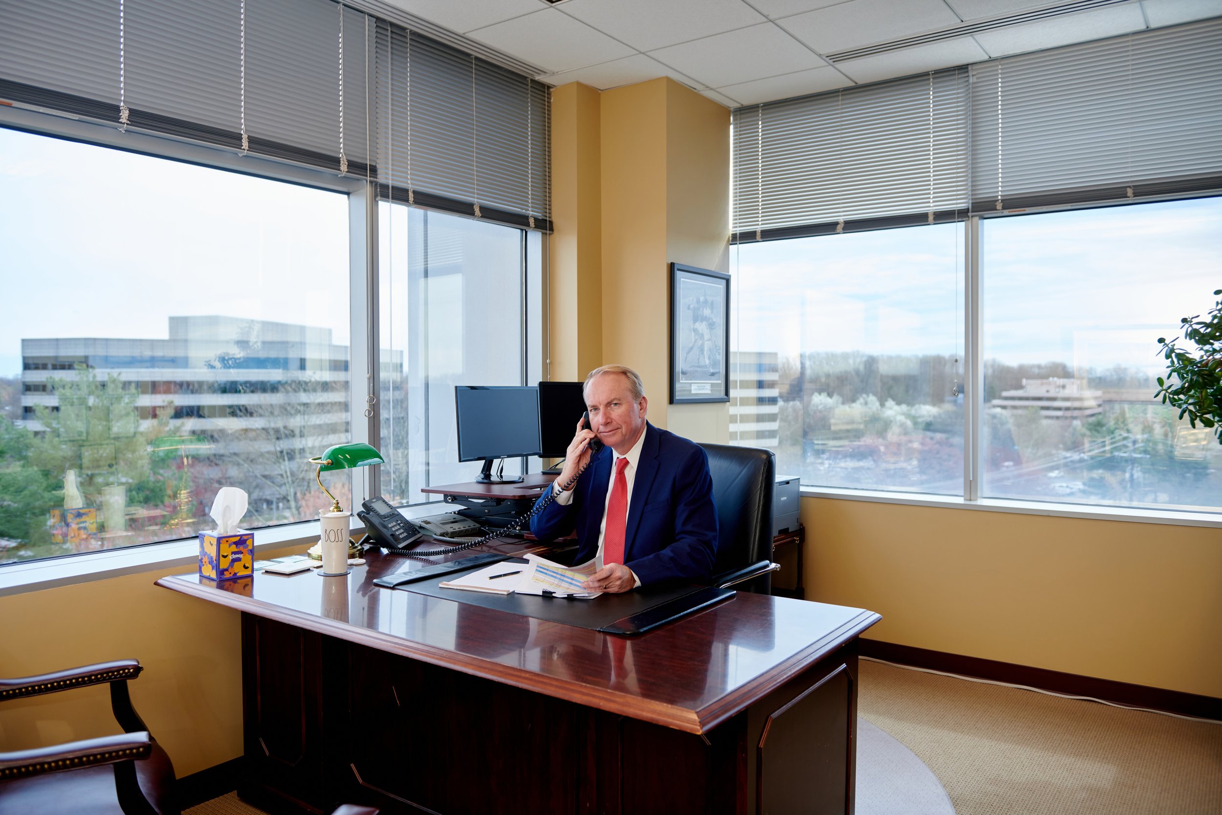 Businessman sitting at desk in modern office talking on phone with window view of cityscape behind him.