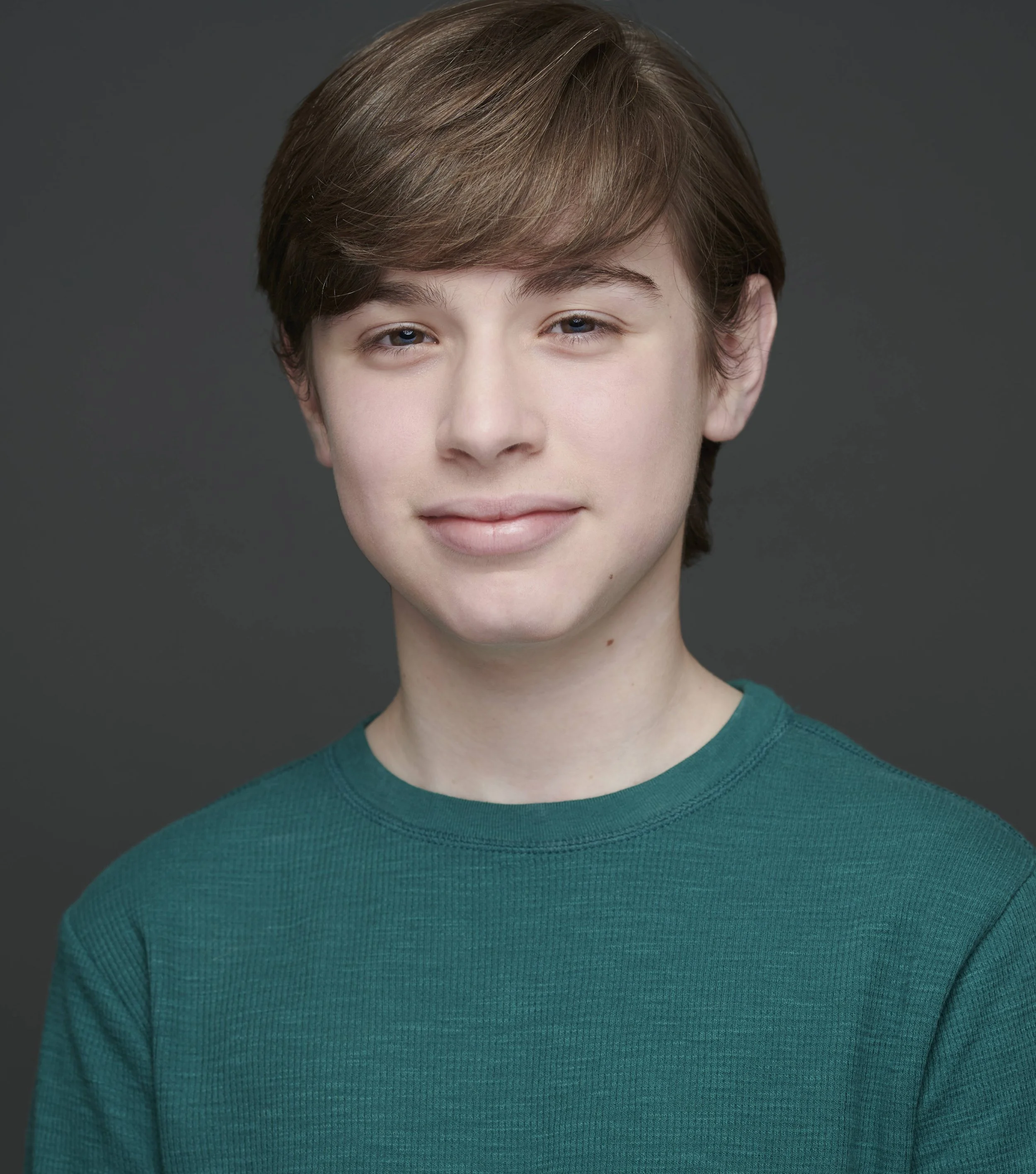 A young person with short, light brown hair wearing a green shirt, smiling softly in front of a gray background.