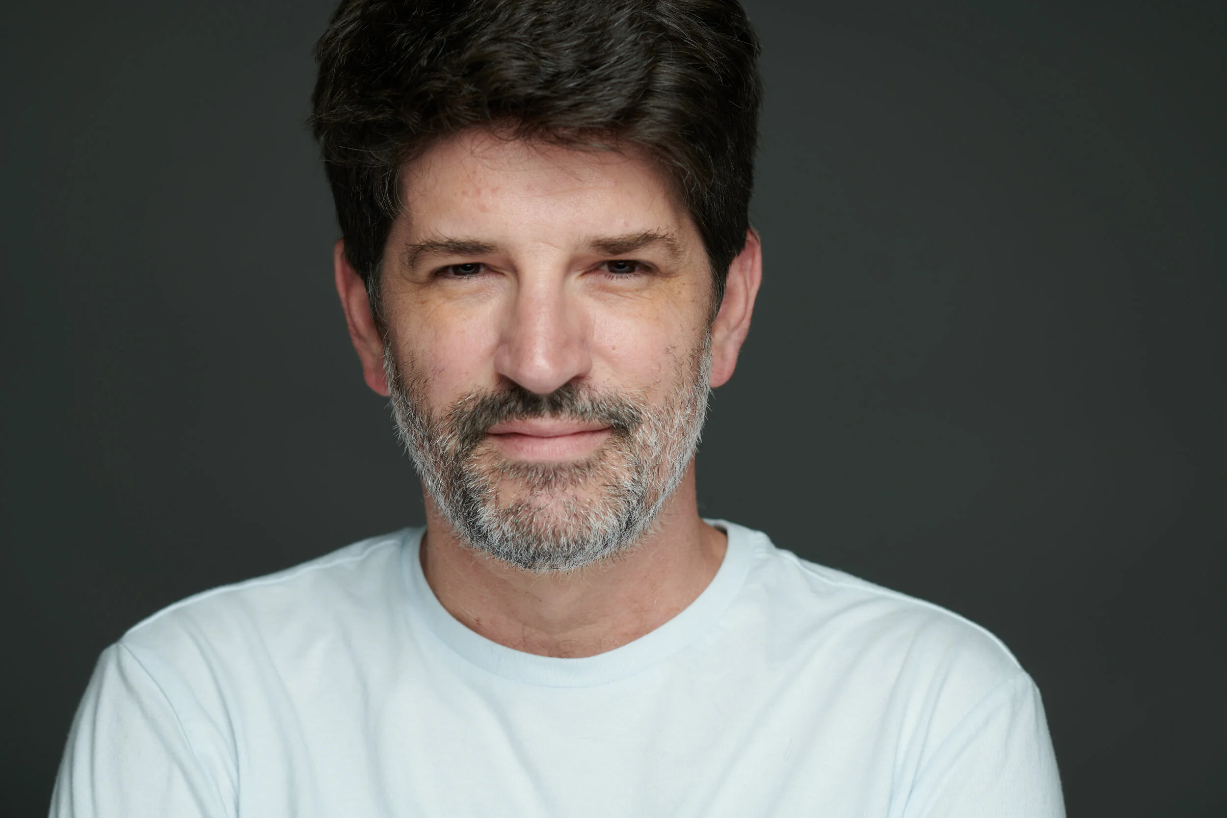 Man with a beard wearing a white T-shirt against a dark background.