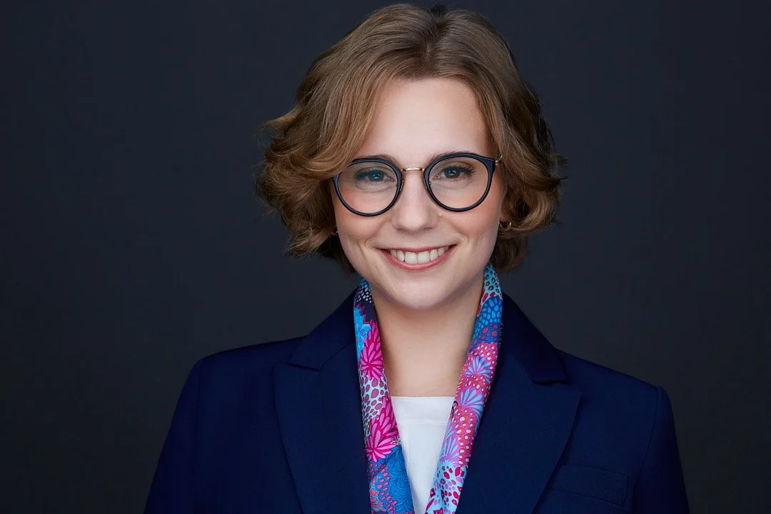 ERAS headshot of a smiling woman with short, wavy blonde hair, wearing glasses, a dark blue blazer, and a colorful scarf against a dark background.
