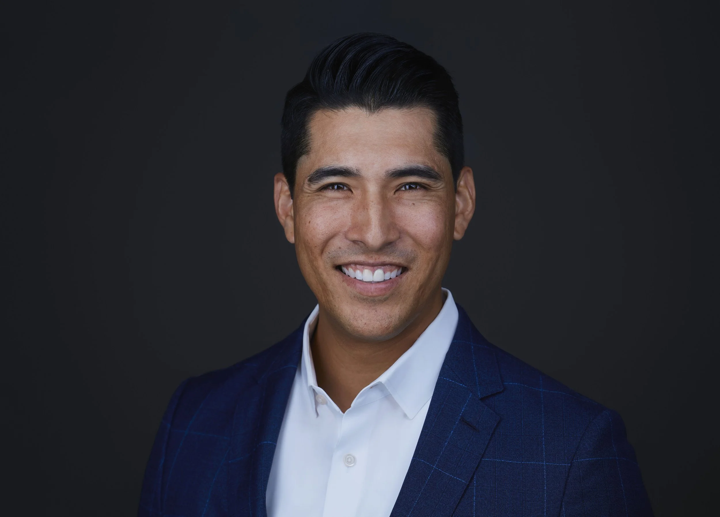 Man in a blue suit smiling against a dark background.