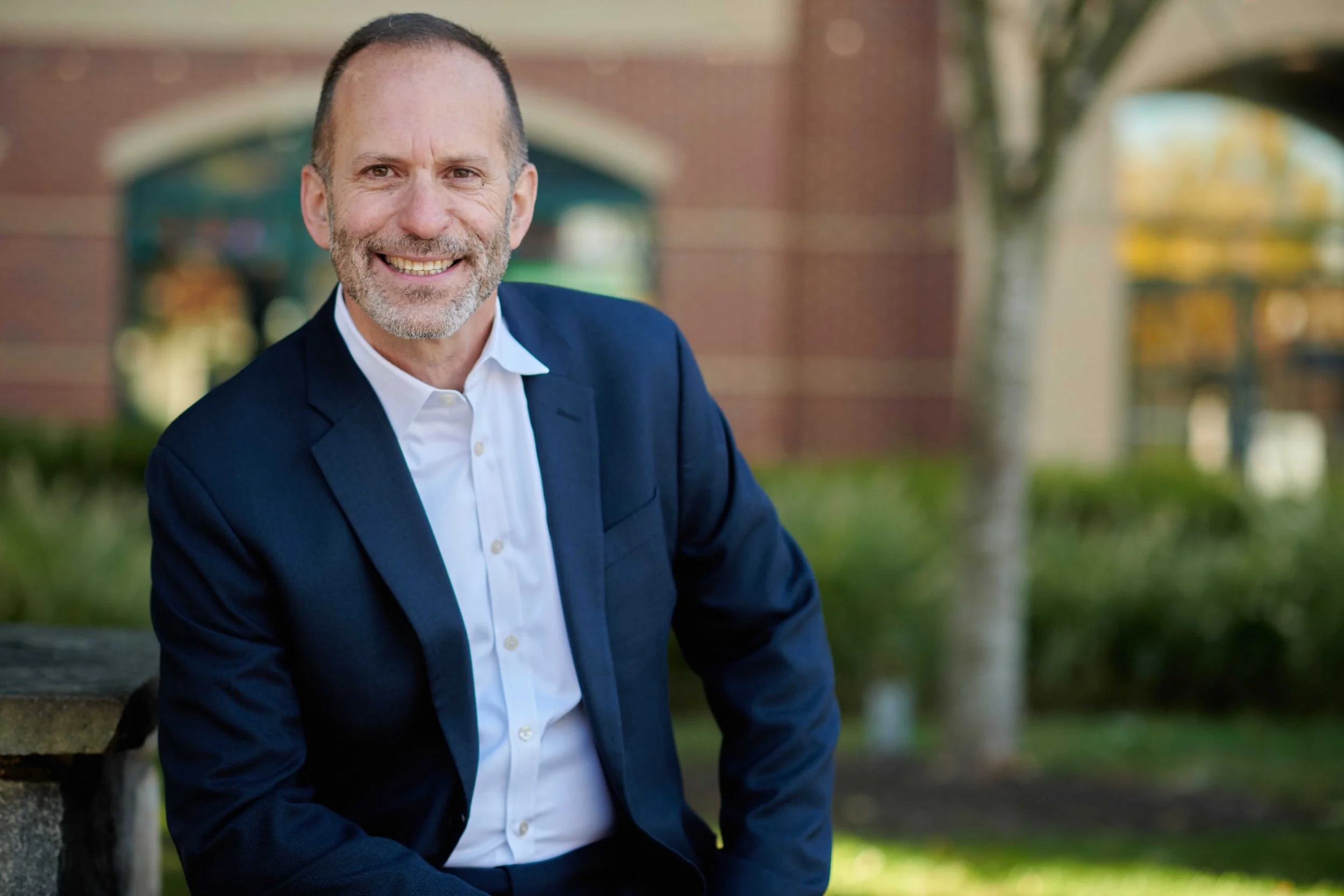 A middle-aged man with short gray hair, beard, and mustache, smiling while wearing a dark blue suit and white shirt, sitting on a park bench outdoors with trees and a brick building in the background.