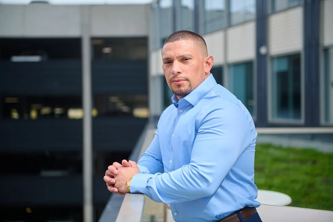 Headshot of a man in a blue dress shirt with his hands clasped, standing outside near a modern building.