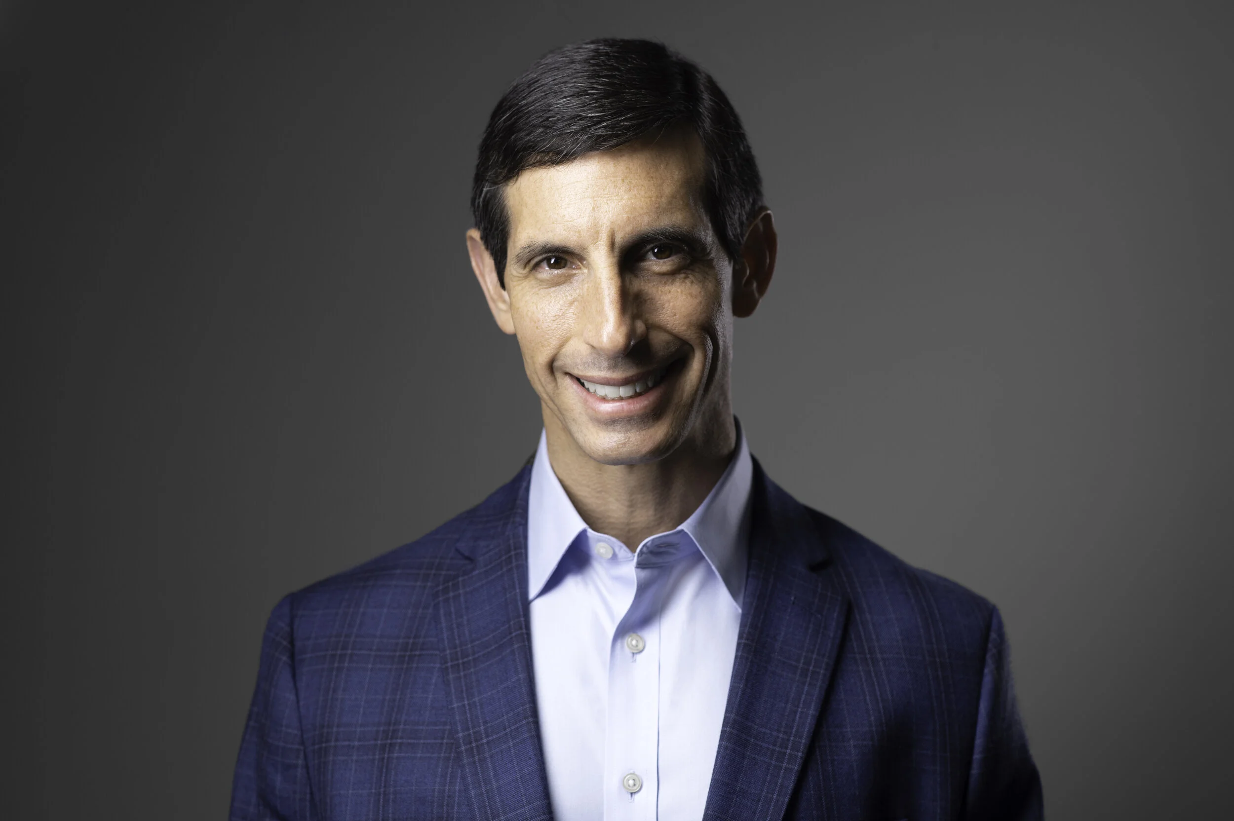 Smiling man in a blue plaid suit jacket and white shirt against a gray background.