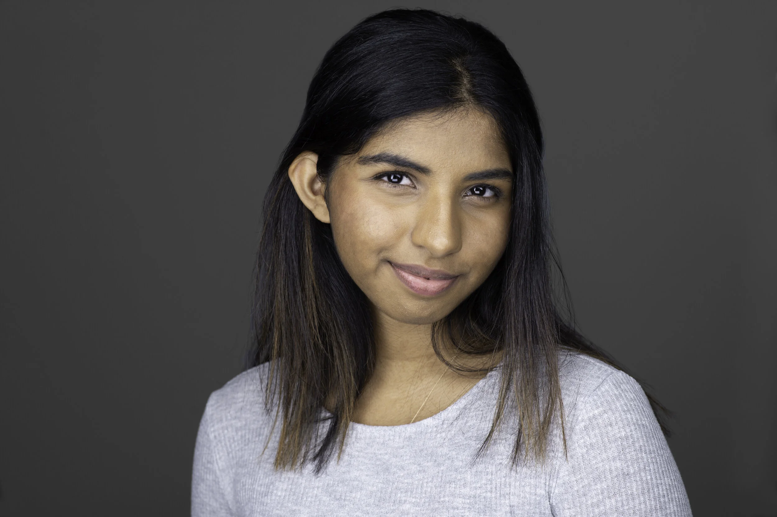 A woman with dark hair wearing a light gray sweater smiles softly in a portrait against a dark gray background.