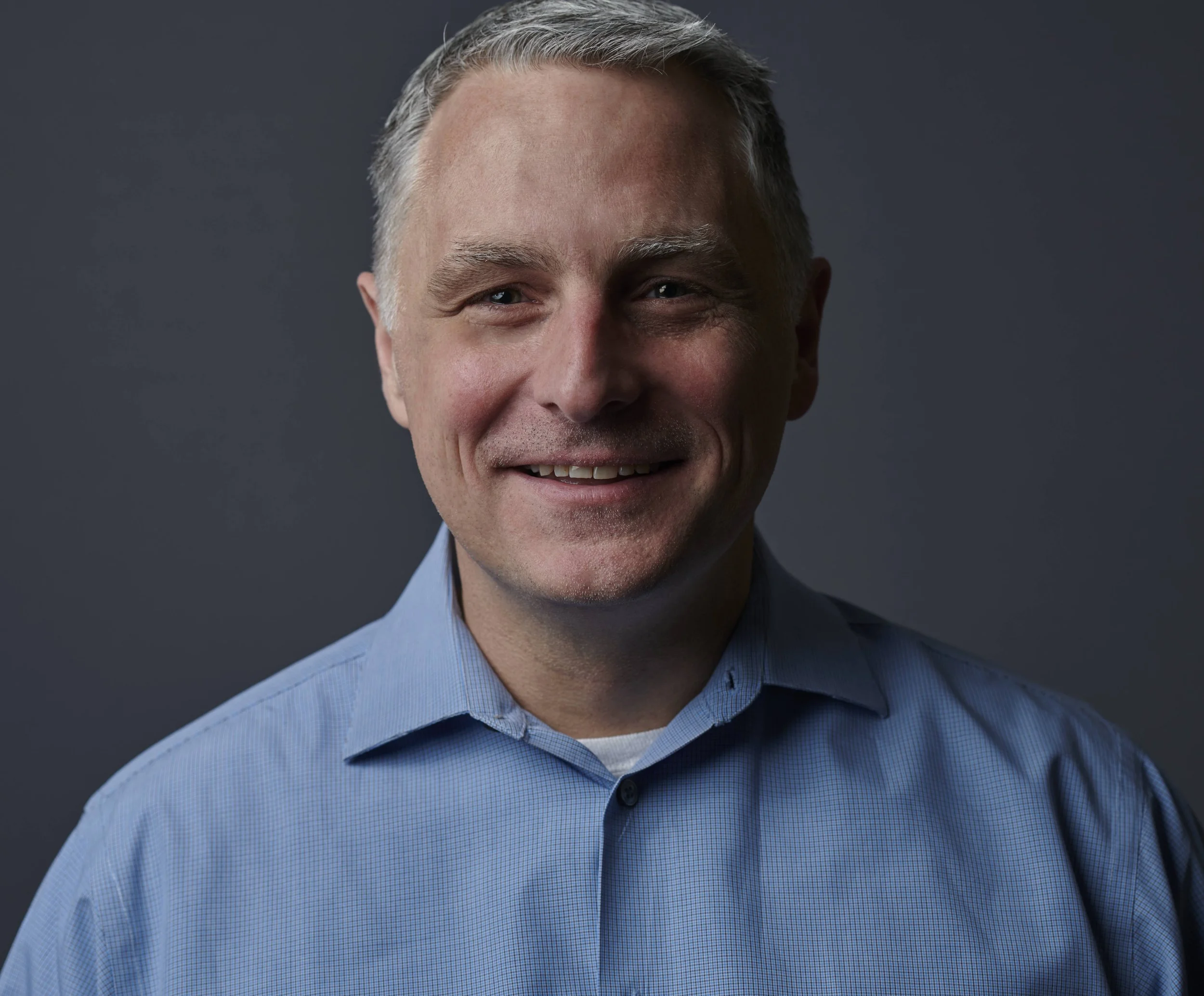Smiling person wearing a blue collared shirt against a dark background.