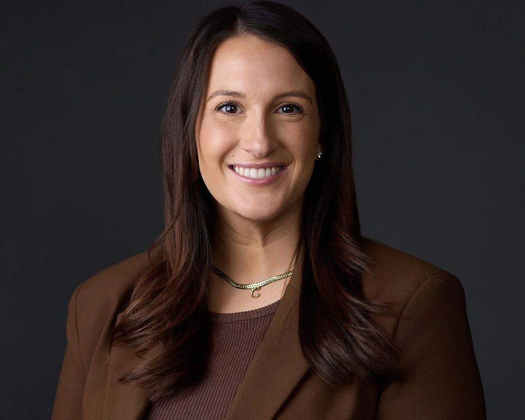 Linkedin Headshot of a  woman with long brown hair smiling against a dark background, wearing a brown blazer and gold necklace.