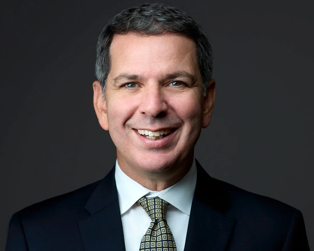 Linkedin Headshot of a smiling man in a dark suit, white shirt, and patterned tie, against a dark background.