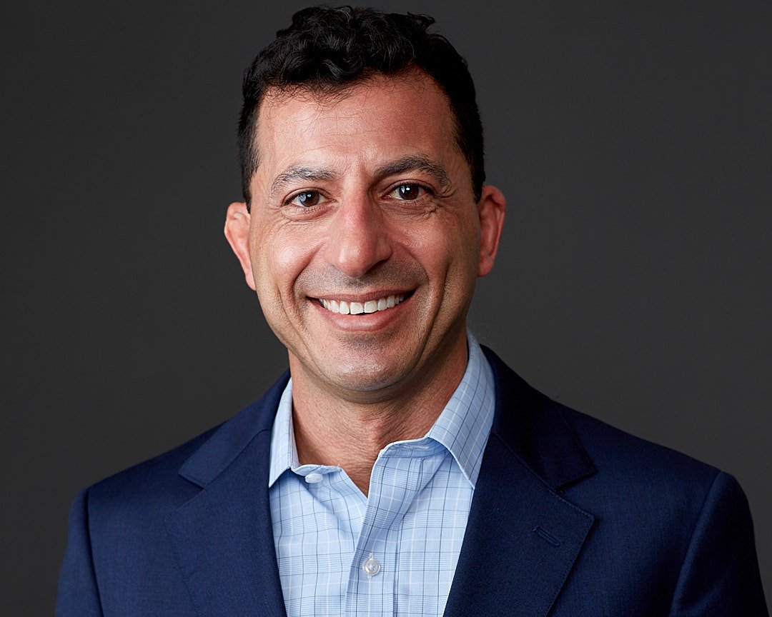 Headshot of a smiling man in a blue suit and light blue dress shirt against a dark background.