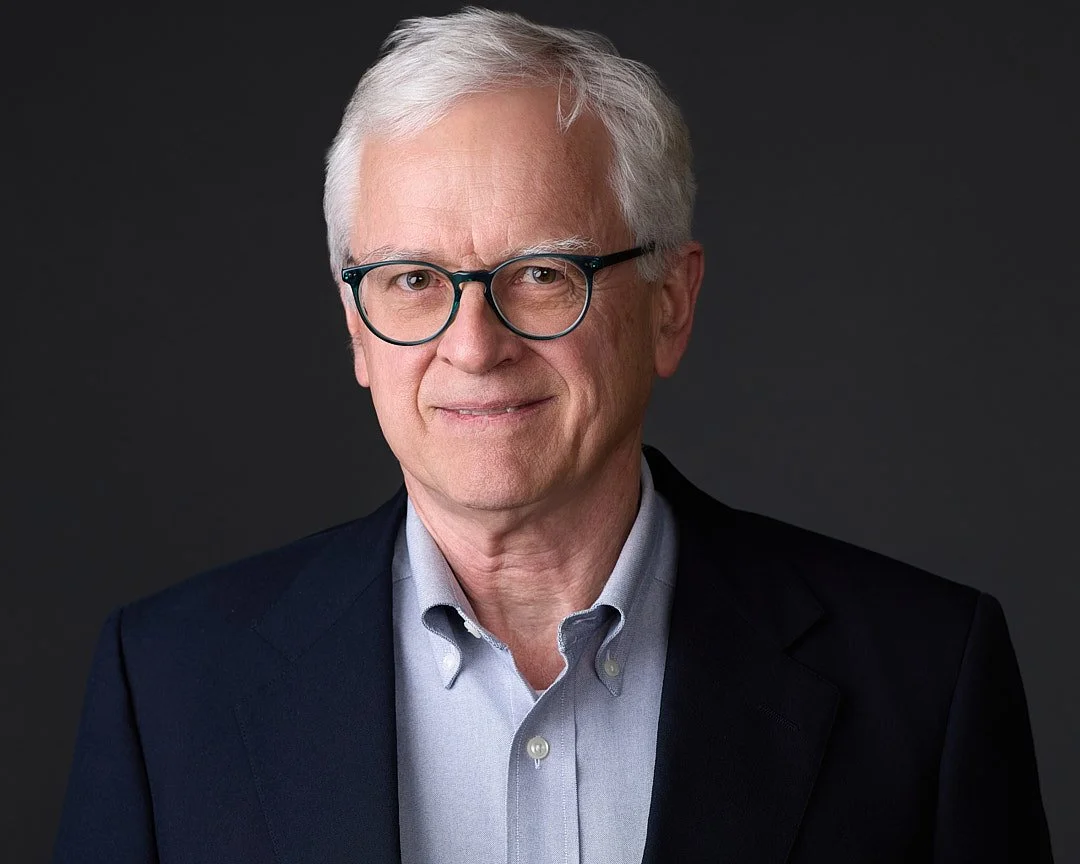 A portrait of an older man with white hair, glasses, and a friendly expression, dressed in a dark suit jacket and light shirt against a dark background.