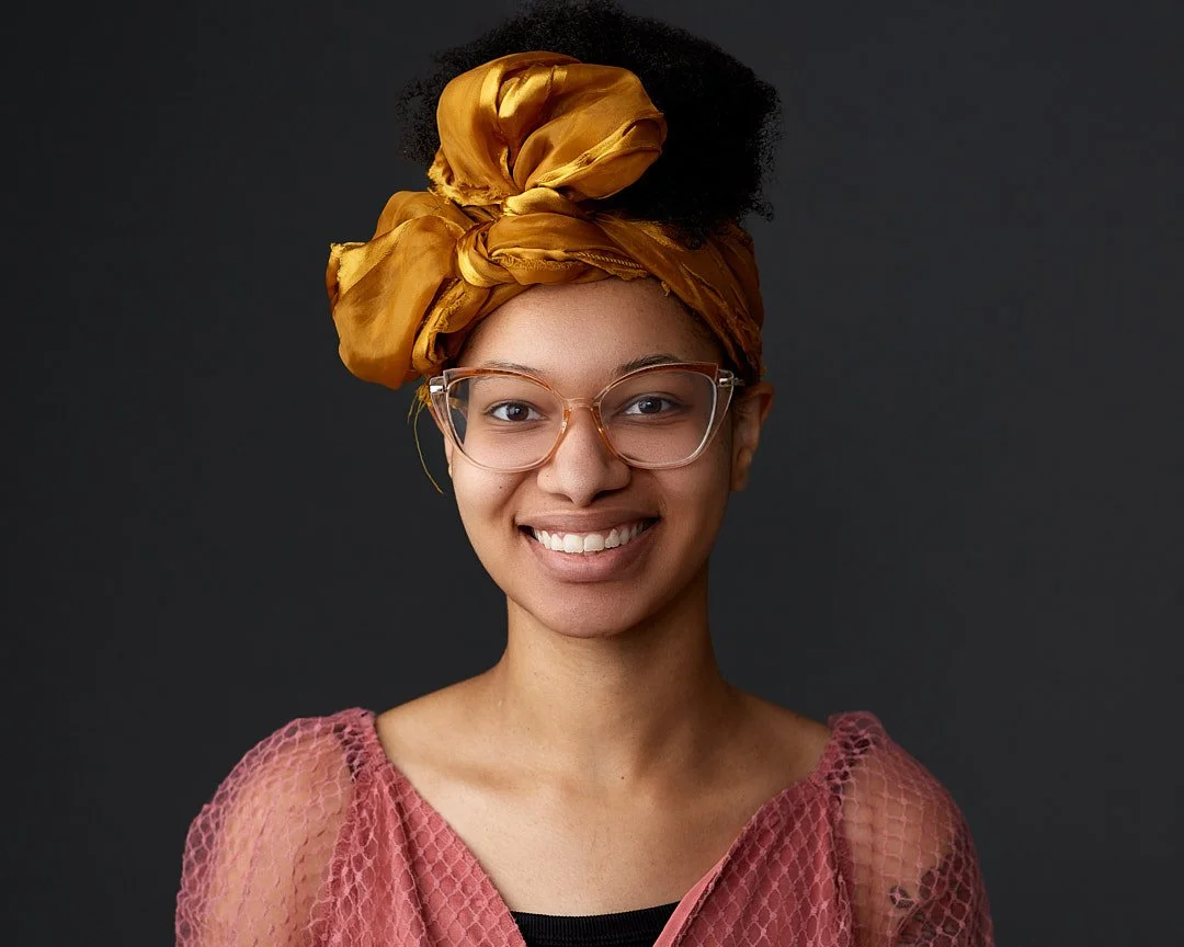 headshot of a young woman with glasses, wearing a pink lace top and a large yellow headwrap, smiling against a dark background.