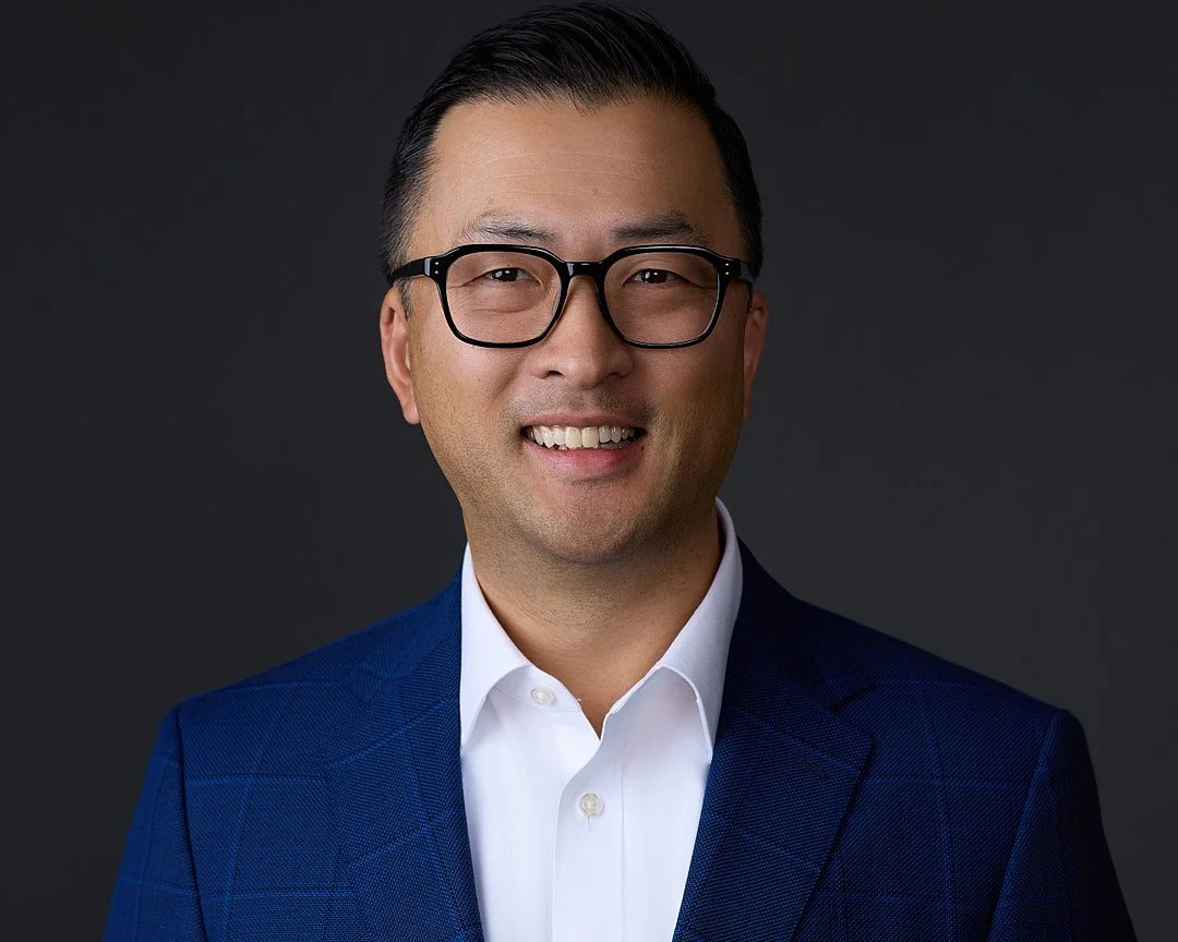 Portrait of a smiling man with black glasses, wearing a navy blue suit and white shirt against a dark background.