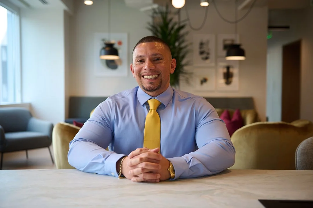 personal branding portrait of A smiling man in a business casual setting, sitting at a table with hands clasped, wearing a light blue shirt and a yellow tie.