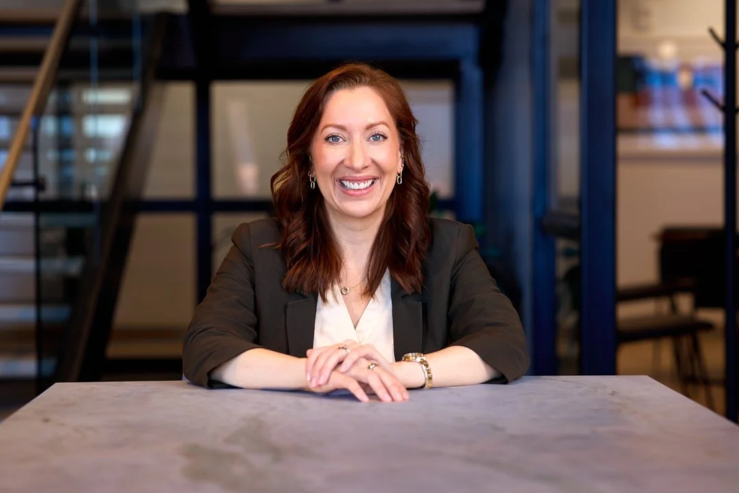personal branding portrait of A woman with red hair, smiling, sitting at a table in an office setting.
