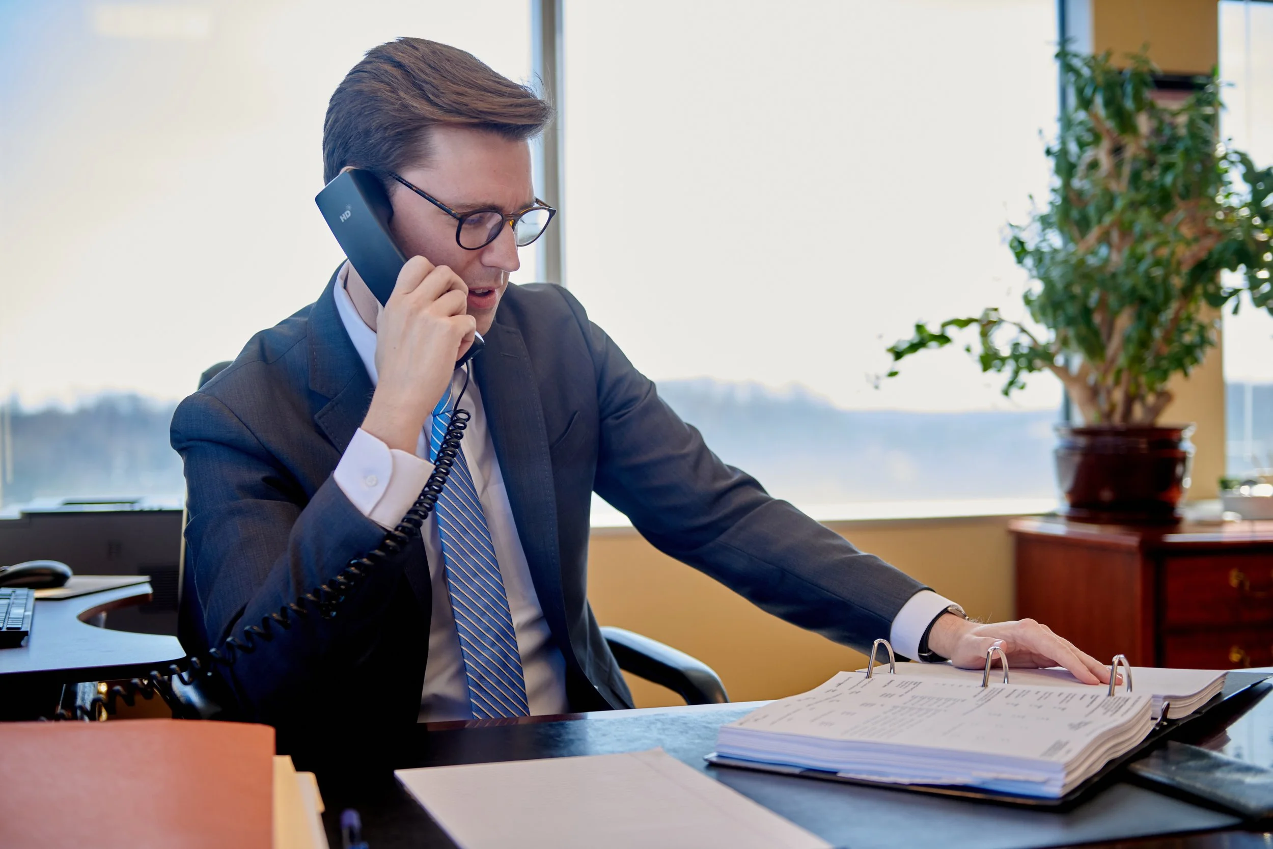 personal branding portrait of A man in a business suit and glasses talking on a landline phone in an office, with documents and a computer on the desk, and a potted plant in the background.