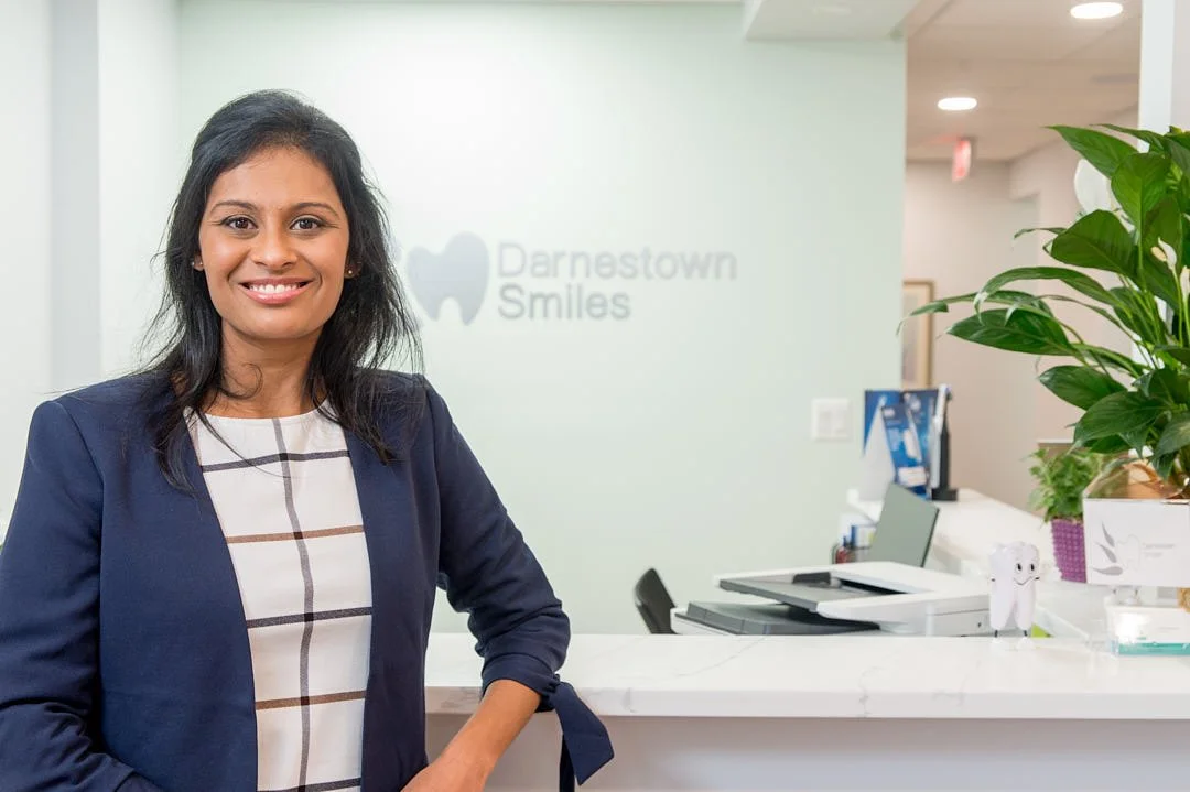 personal branding portrait of A woman standing at a reception desk in a dental office, smiling, with a sign that reads "Darnestown Smiles" in the background.