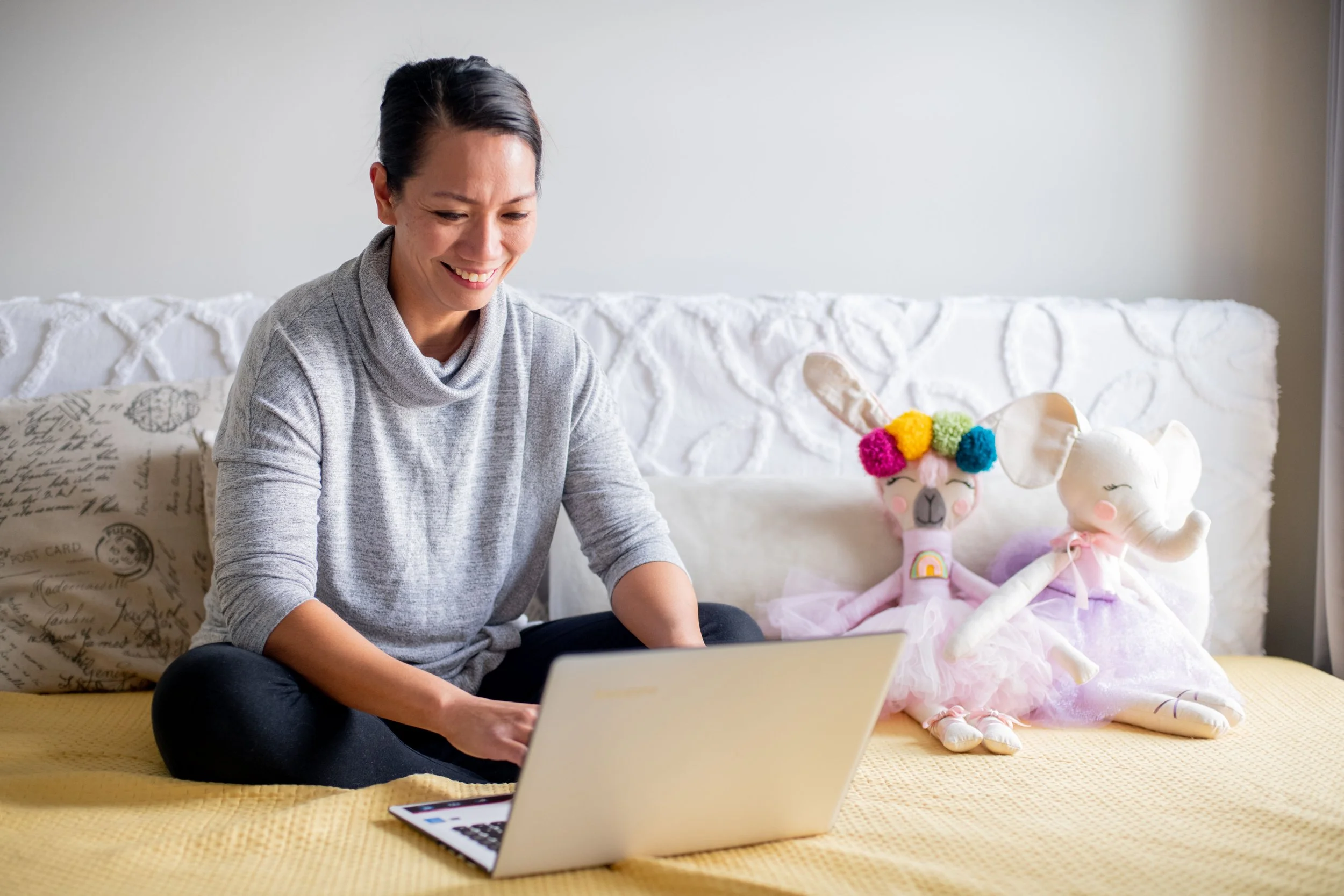 personal branding portrait of A woman sitting on a bed using a laptop, with two stuffed animal toys, a unicorn and a llama, sitting next to her.