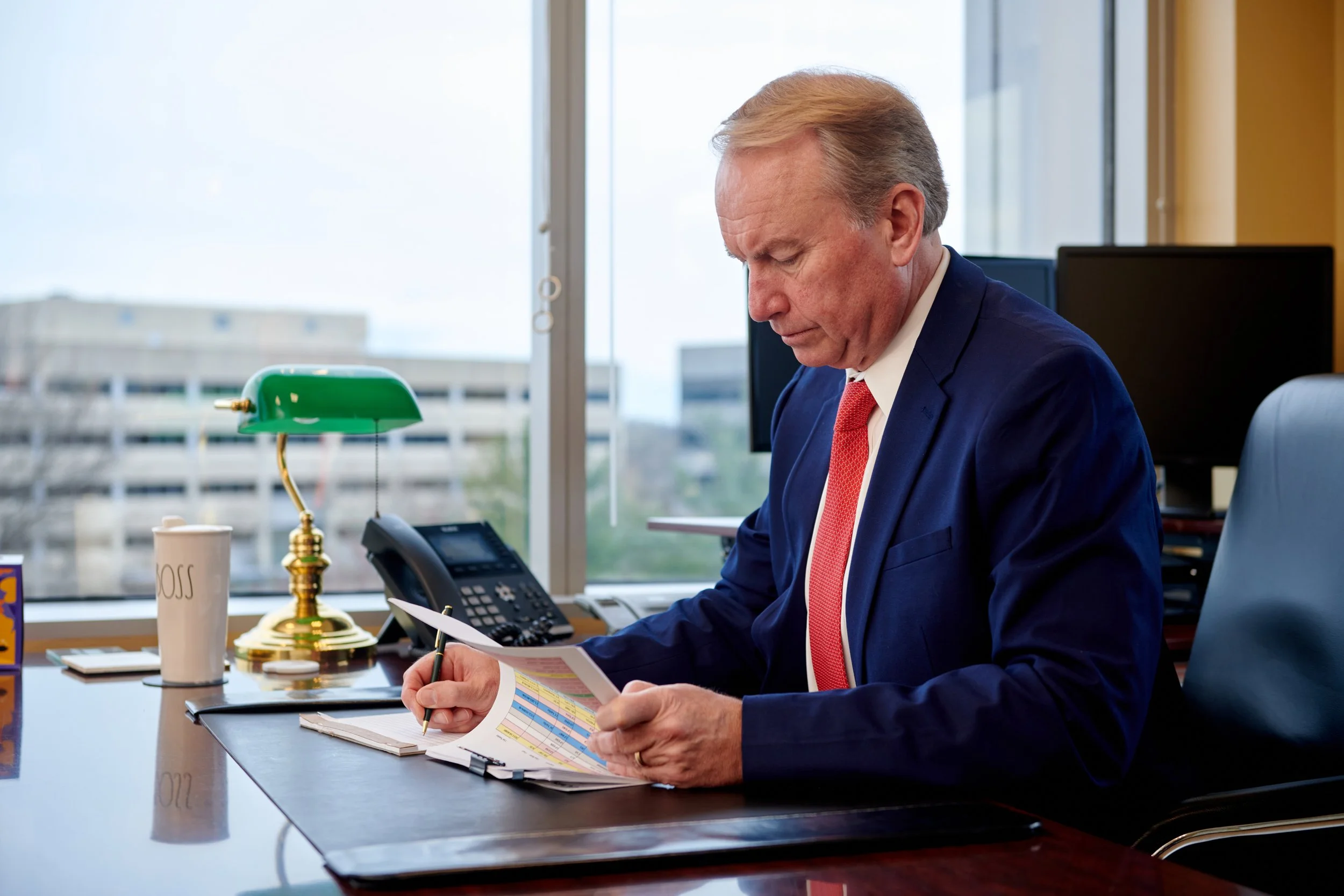 personal branding portrait of A businessman in a blue suit and red tie sitting at a desk, reviewing documents with a spreadsheet, in an office with large windows.
