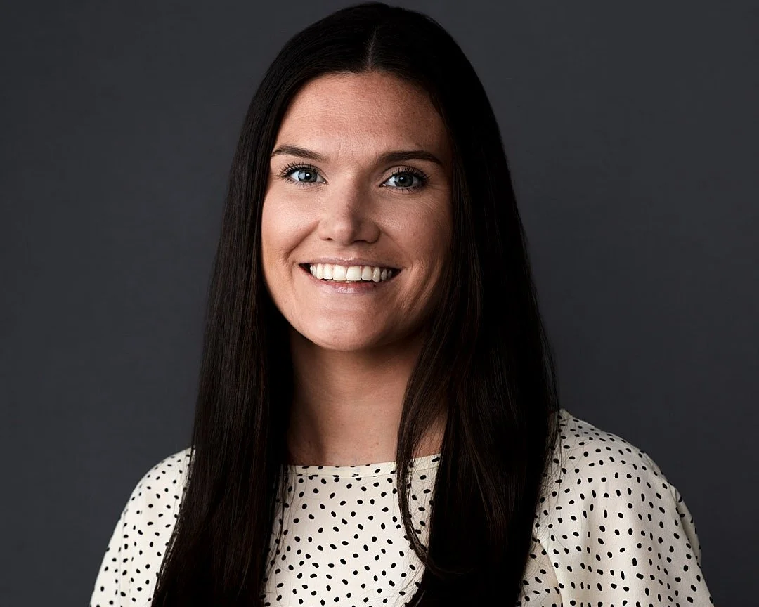 Headshot of a Smiling woman with long dark hair wearing a polka dot top against a dark background.