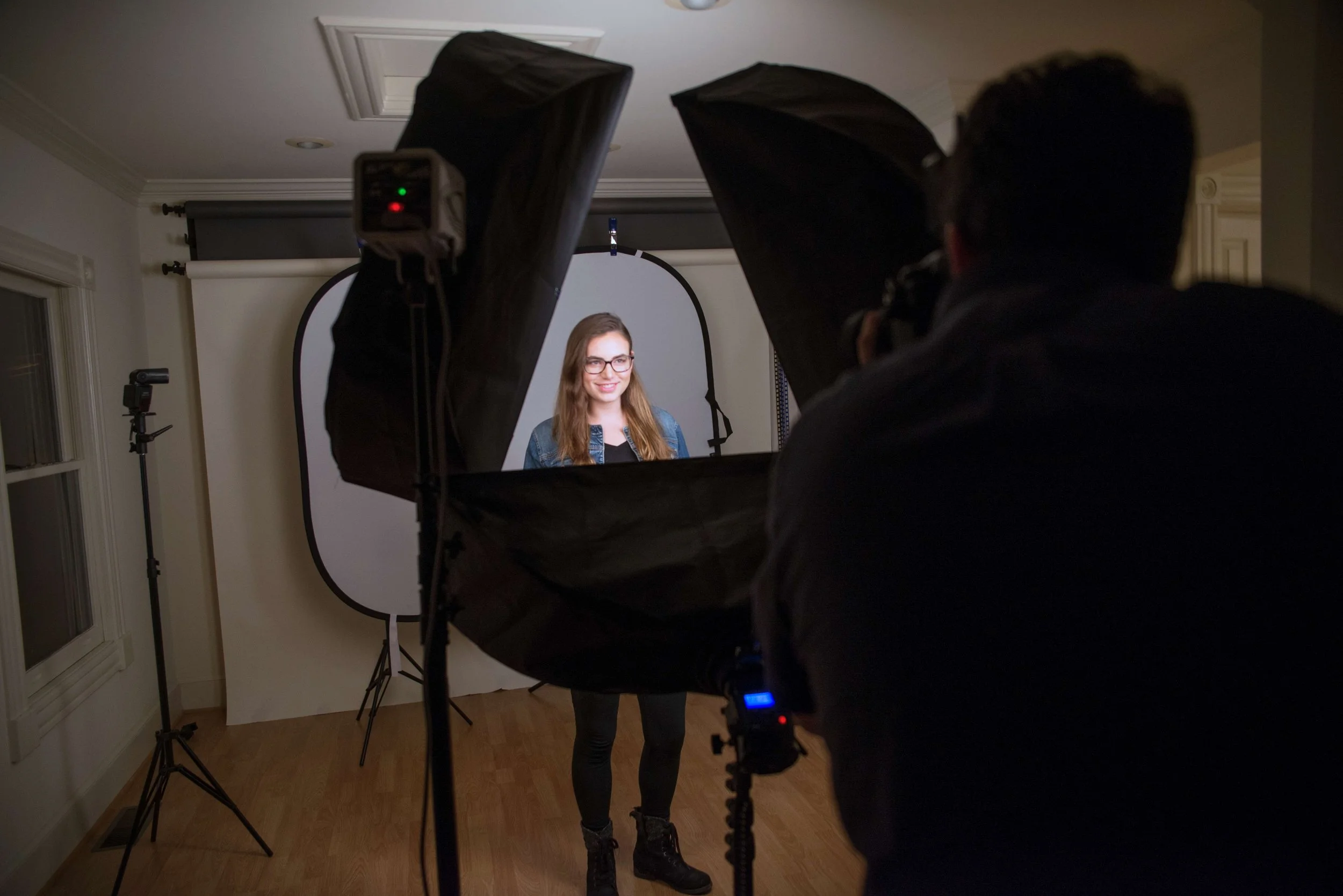 A photographer taking a portrait of a woman in a studio setup with softbox lights and a white backdrop.