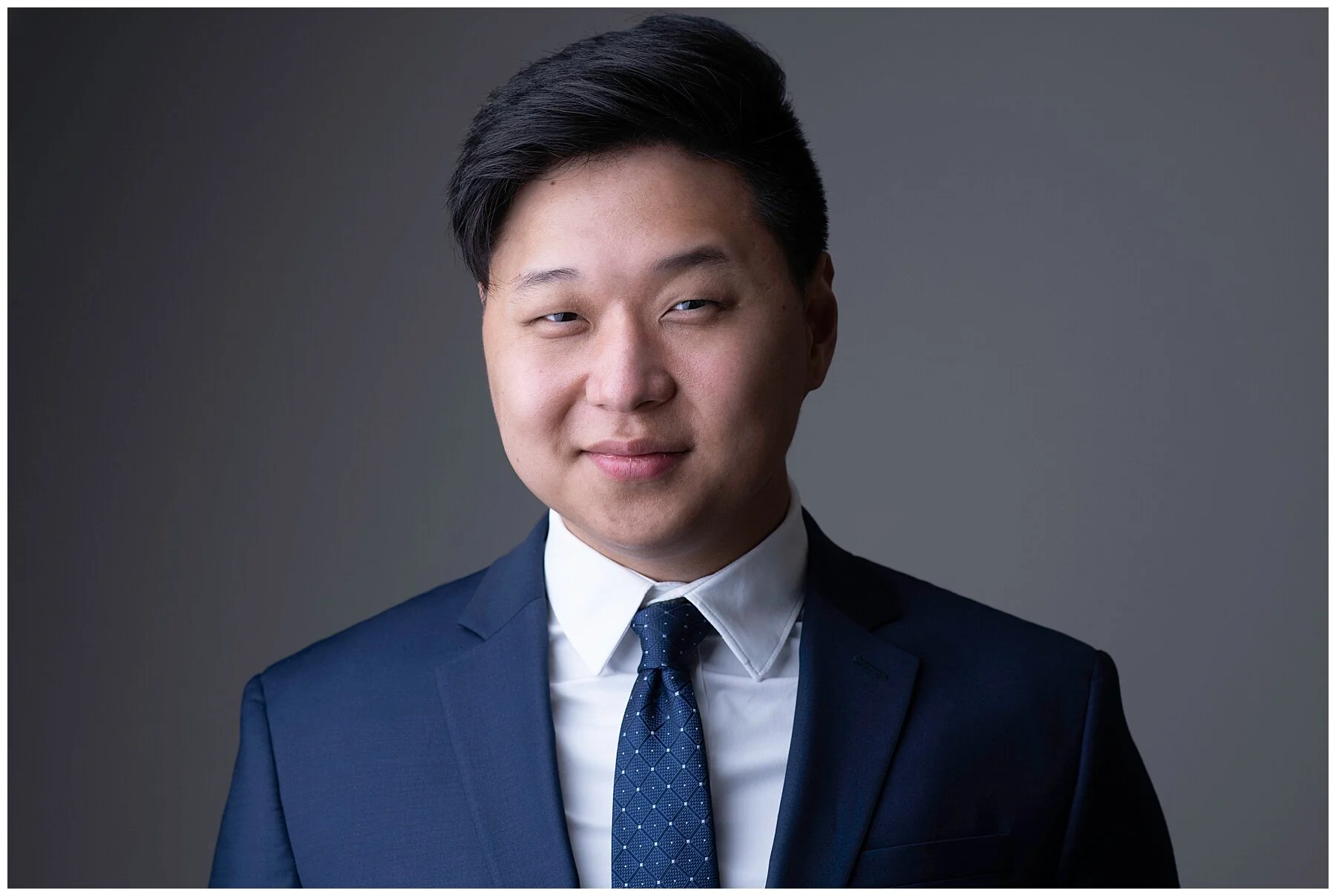 Man in a dark suit and tie with a slight smile, against a gray background.