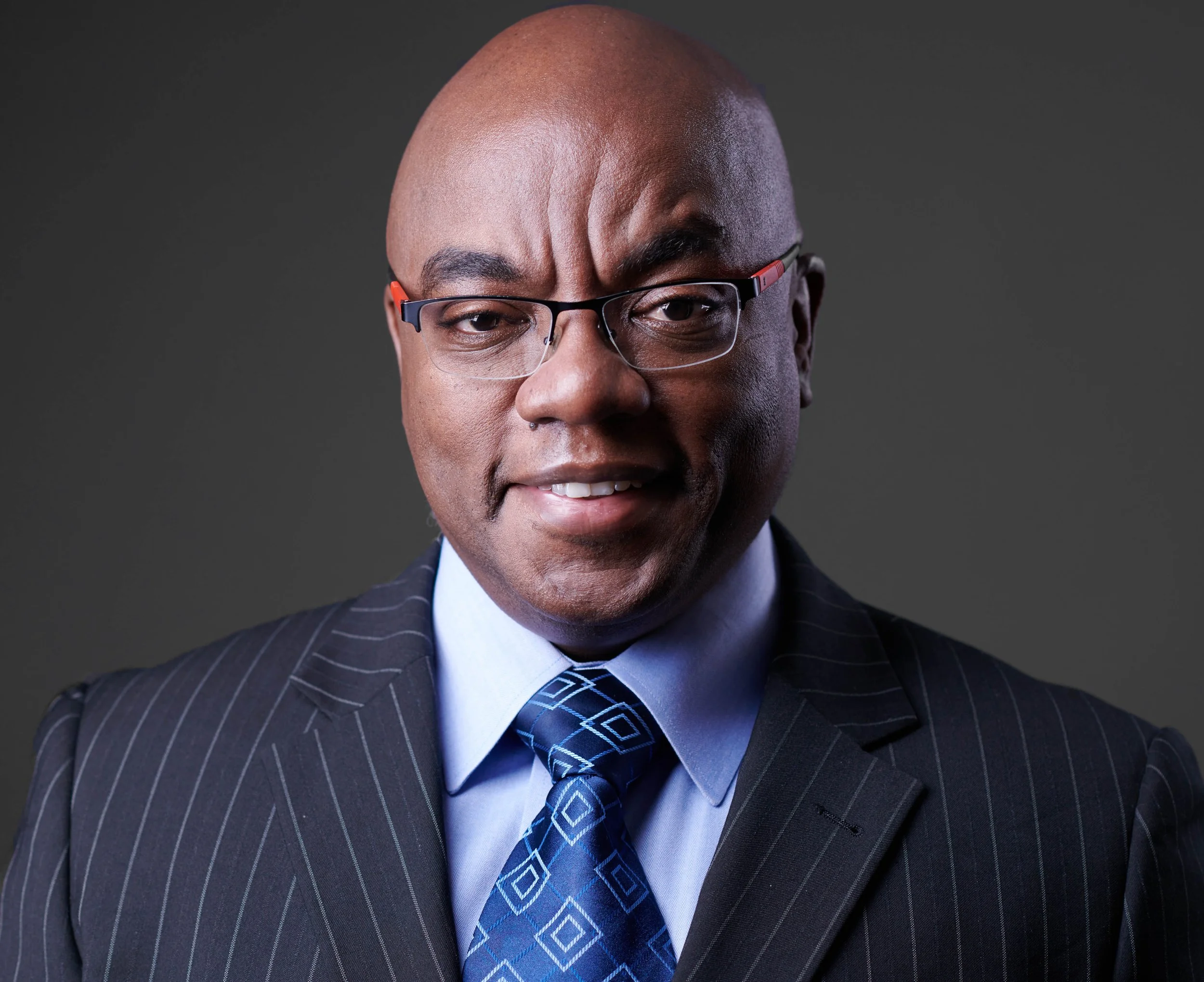 Bald man wearing glasses, a pinstripe suit, and a blue patterned tie, smiling against a gray background.