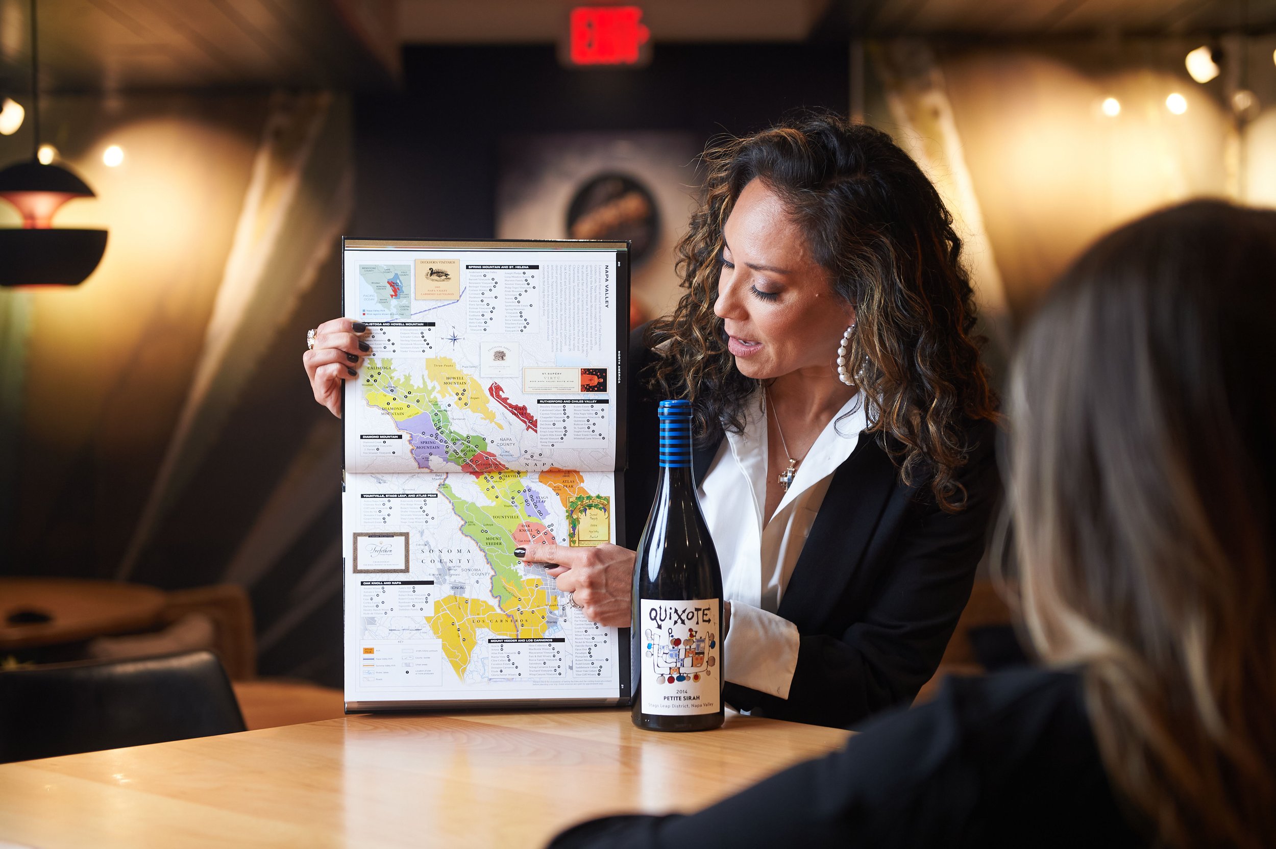 Woman in a black blazer pointing at a colorful Napa Valley wine map, seated at a table with a bottle of Quixote Petite Sirah wine in front.