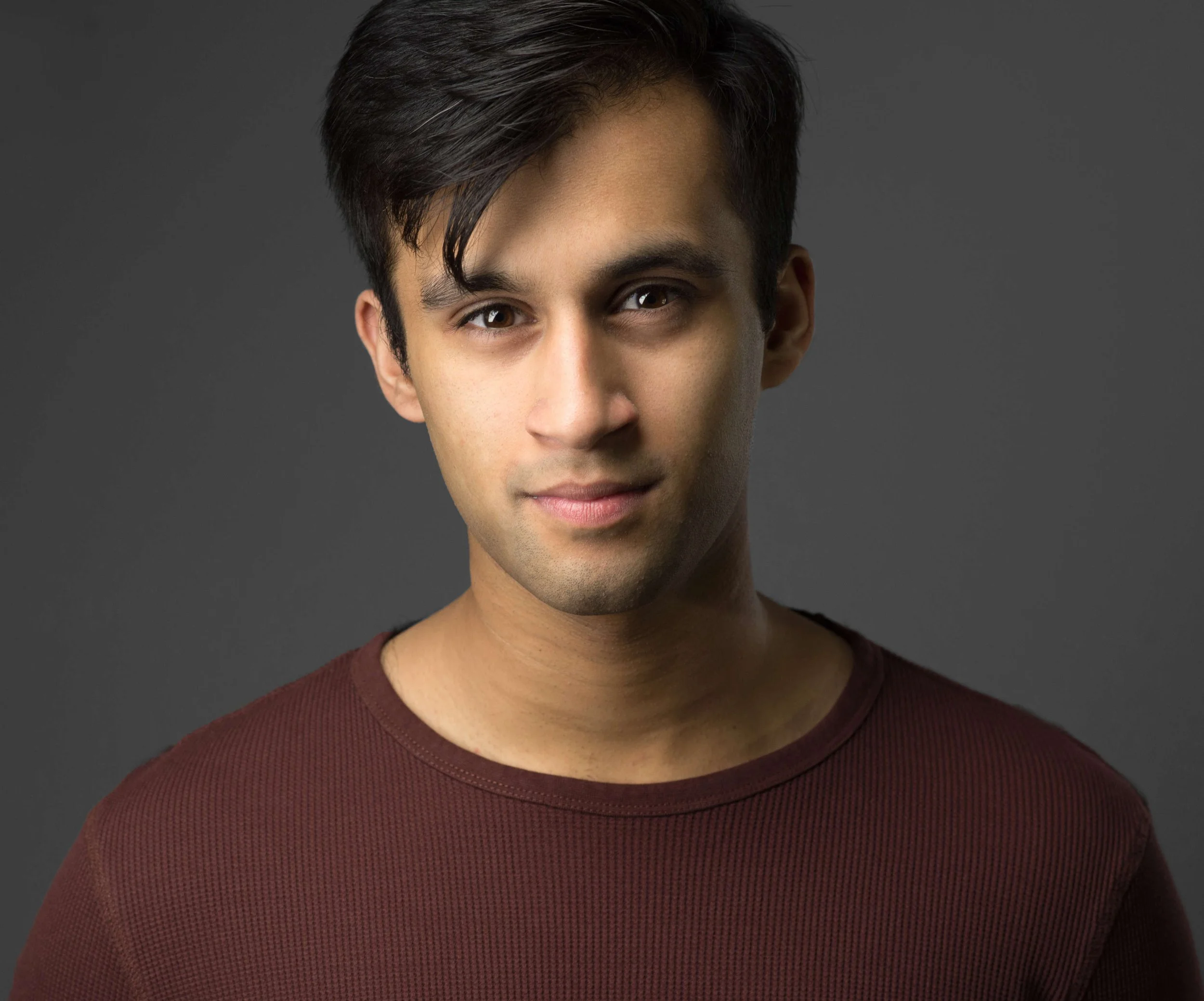A person with short dark hair wearing a dark maroon sweater on a gray background.