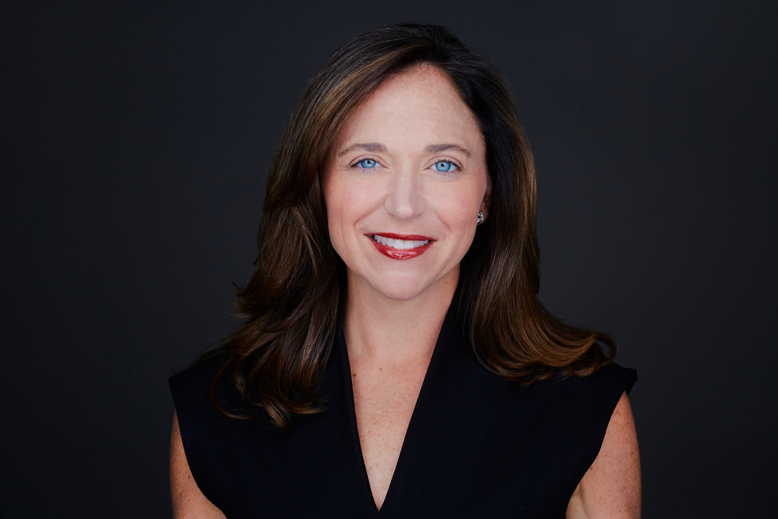 Portrait of a woman with long brown hair and blue eyes, wearing a black top, smiling against a dark background.