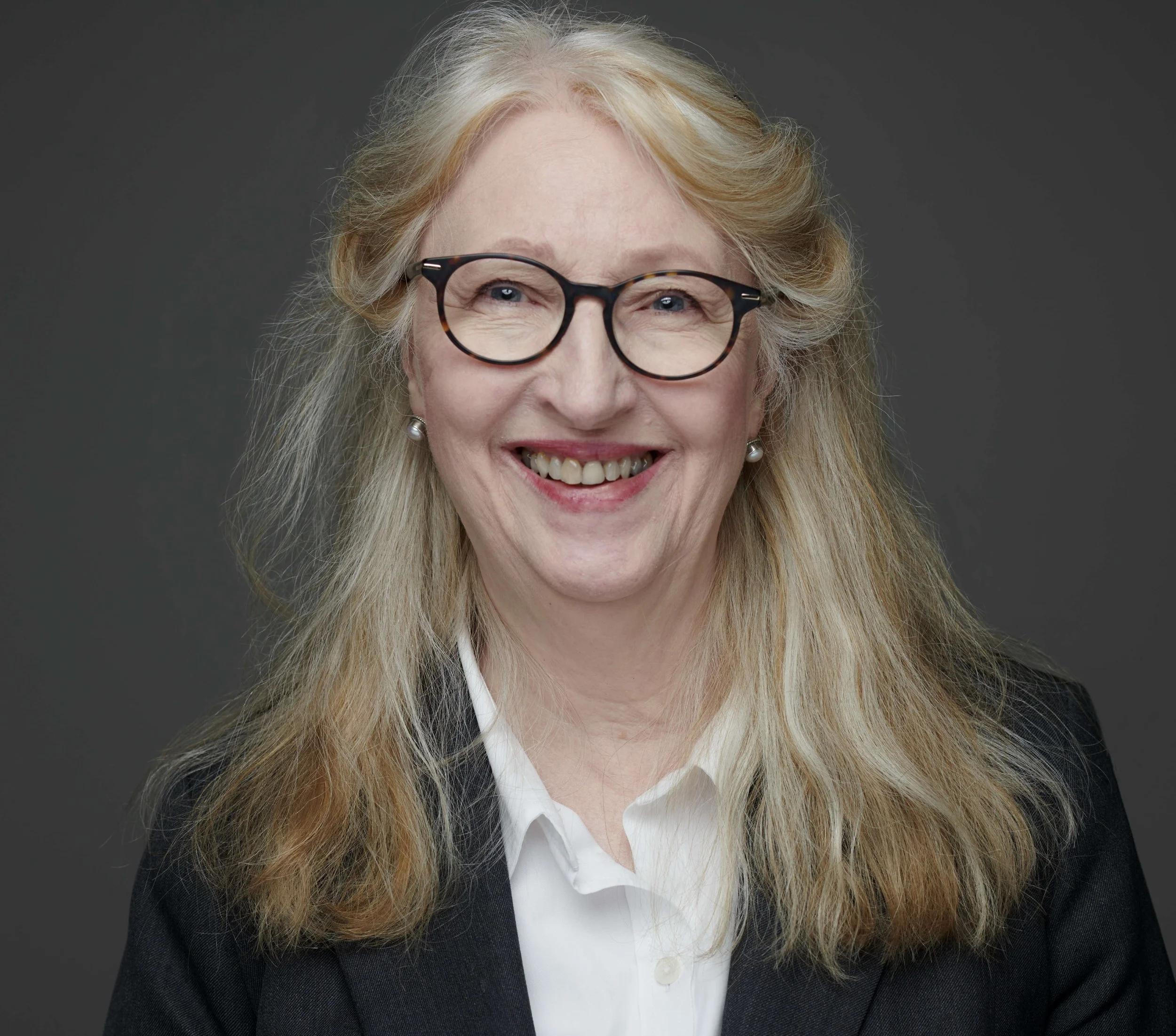 A woman with long blonde hair and glasses, wearing a white shirt and dark blazer, smiling.
