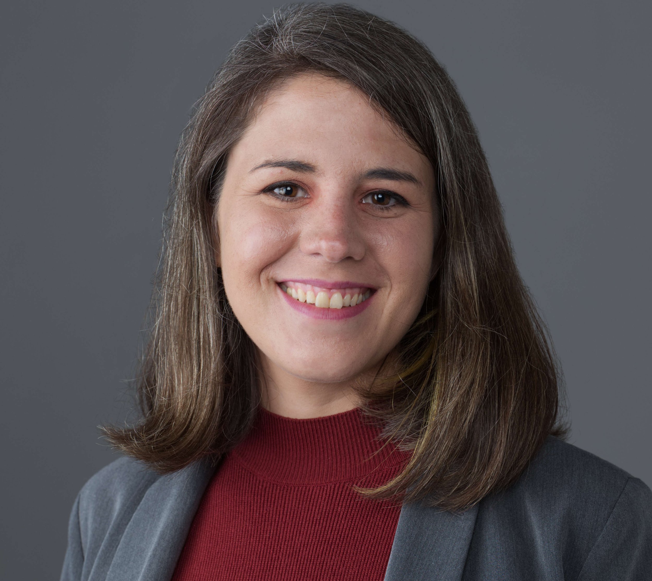 A smiling person with long dark hair wearing a gray blazer and a red sweater against a gray background.
