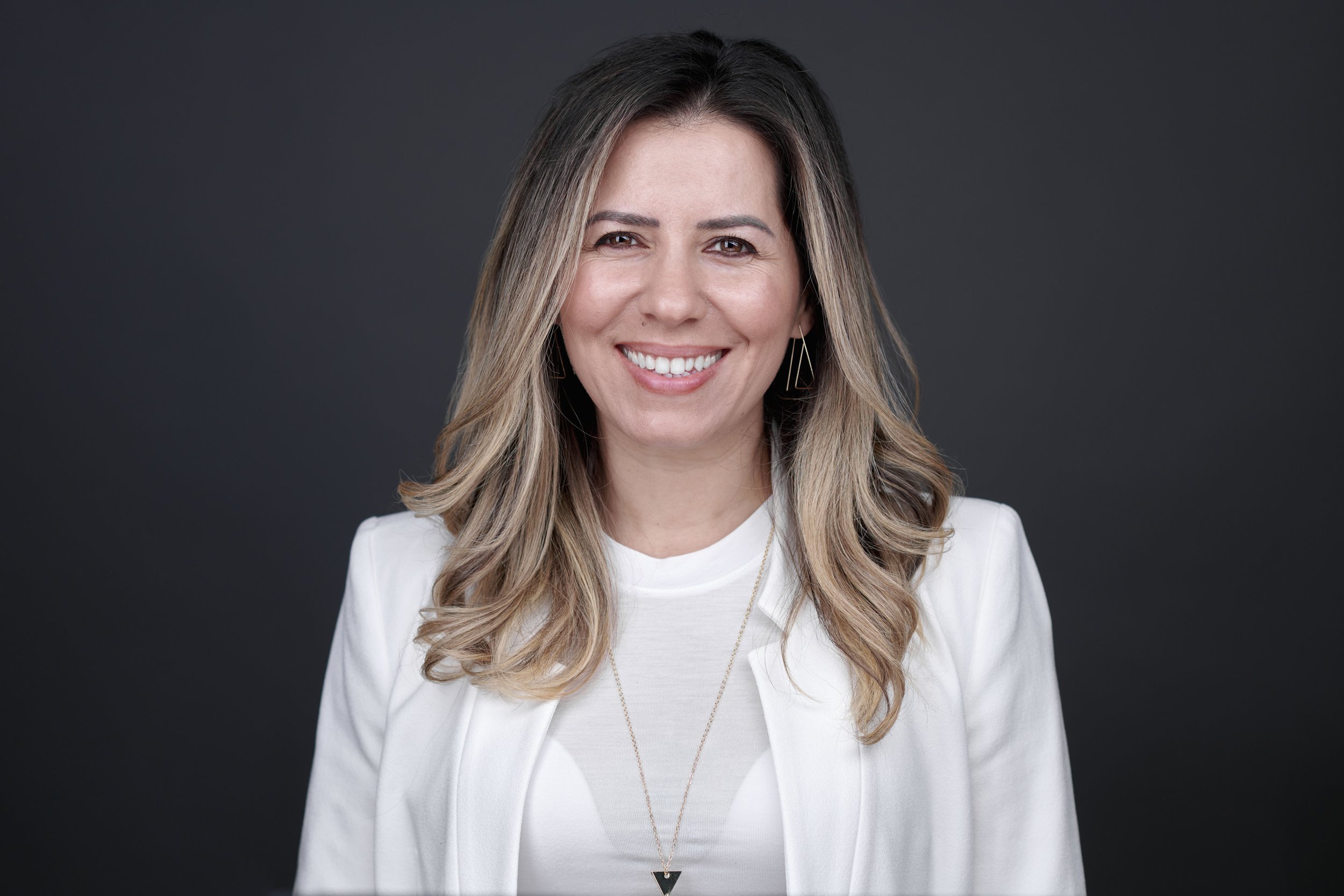 Smiling woman with long hair wearing a white blazer against a dark background.