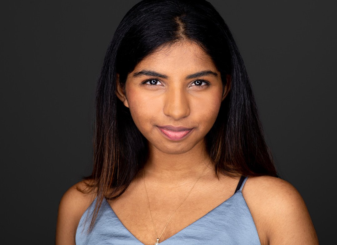 Headshot of a young woman with long dark hair, wearing a light blue tank top and a delicate necklace, against a dark background.