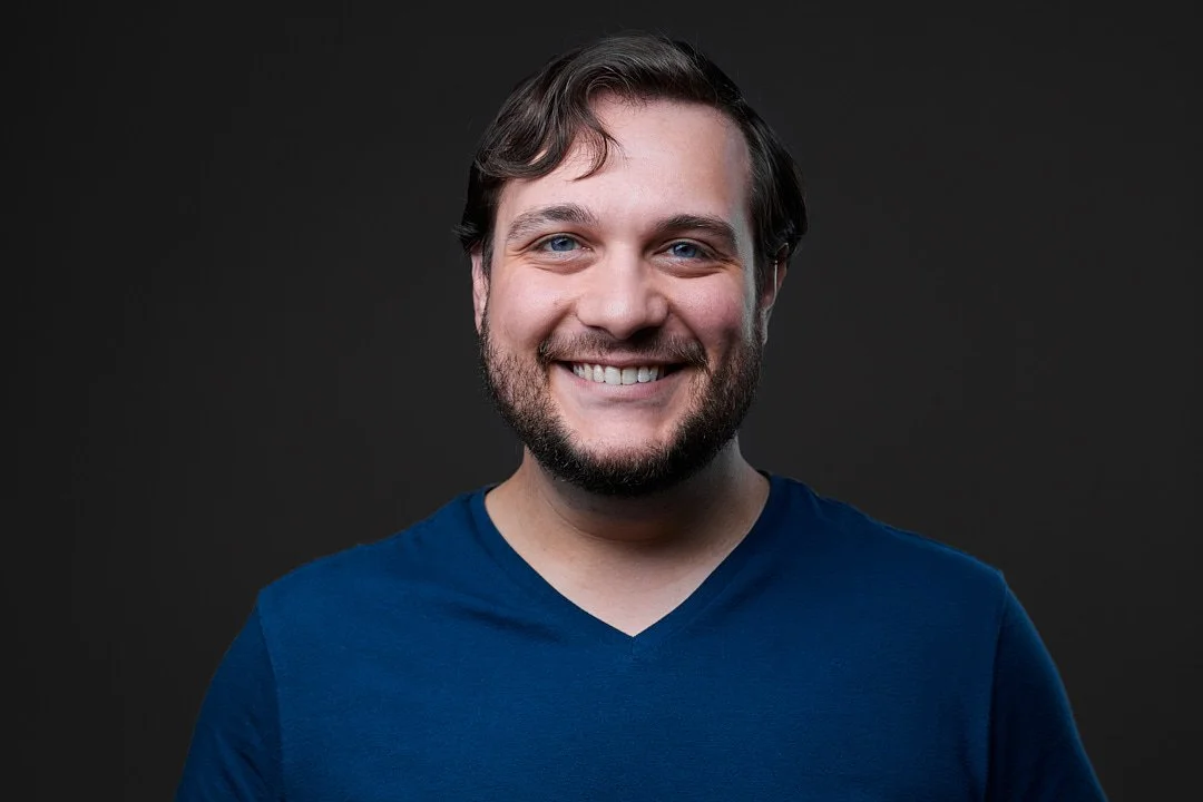 Headshot of a man with short brown hair and beard wearing a blue shirt against a dark background.