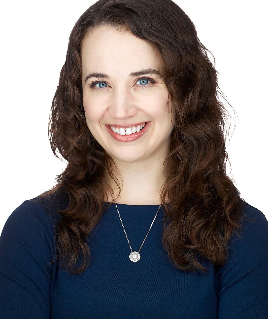Headshot of a  woman with long, wavy brown hair and blue eyes smiling, wearing a dark blue top and a silver necklace, against a white background.
