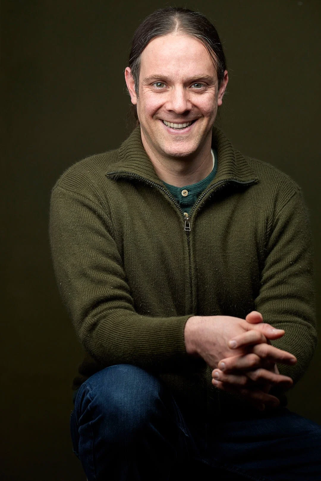 Portrait of a male actor sitting on a stool wearing a green sweater against a green backdrop