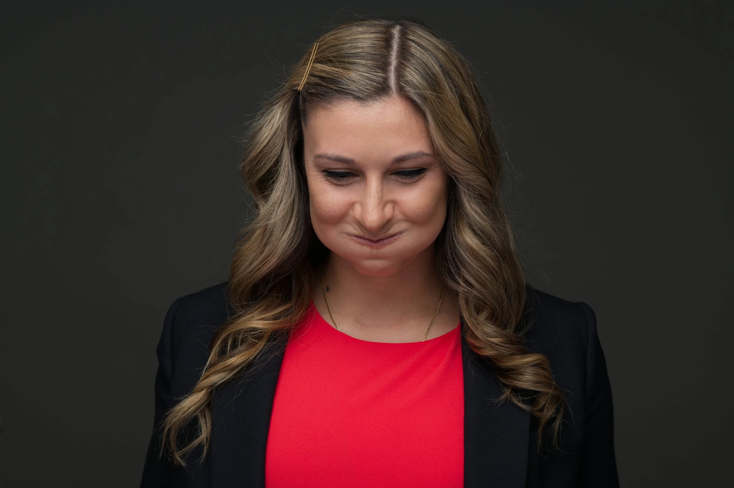 A woman with long wavy hair and a red top smiling with her mouth closed, appearing to puff her cheeks.