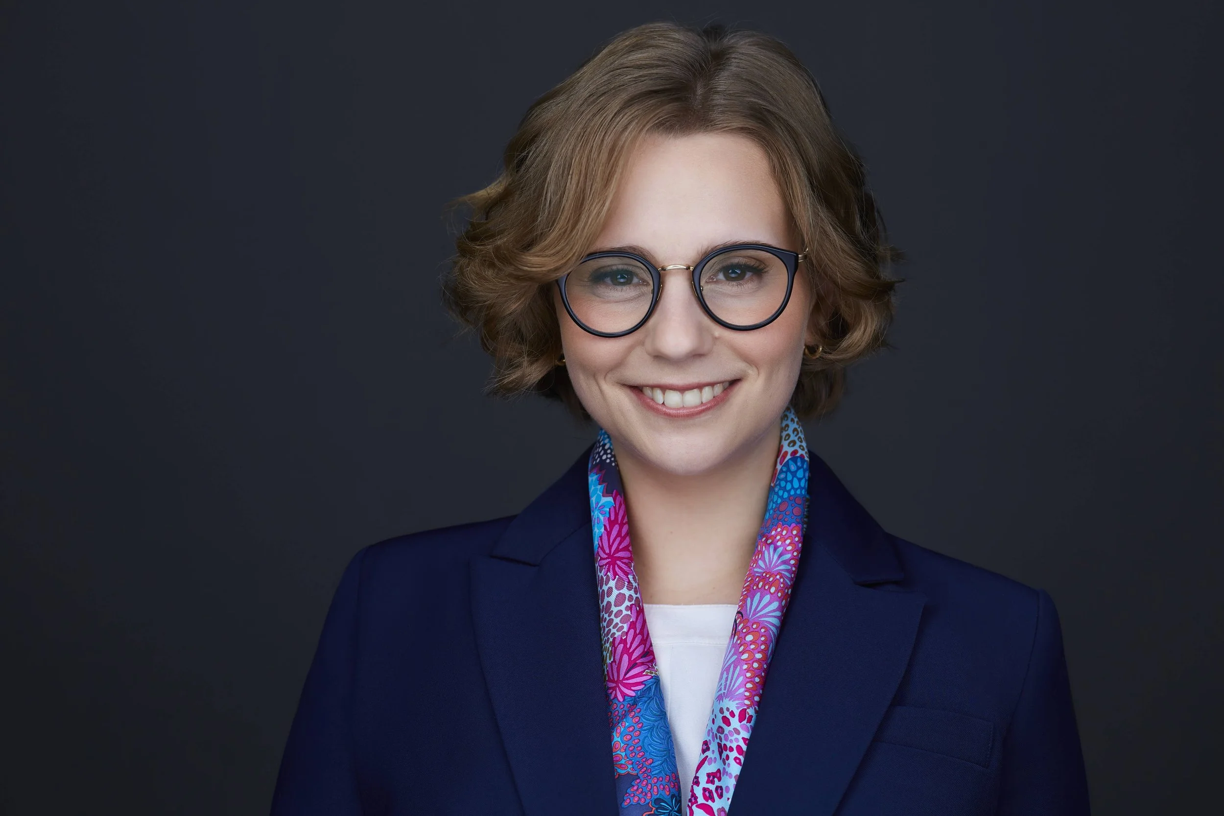 Portrait of a person with short hair and glasses, wearing a colorful scarf and a dark blazer, smiling against a dark background.
