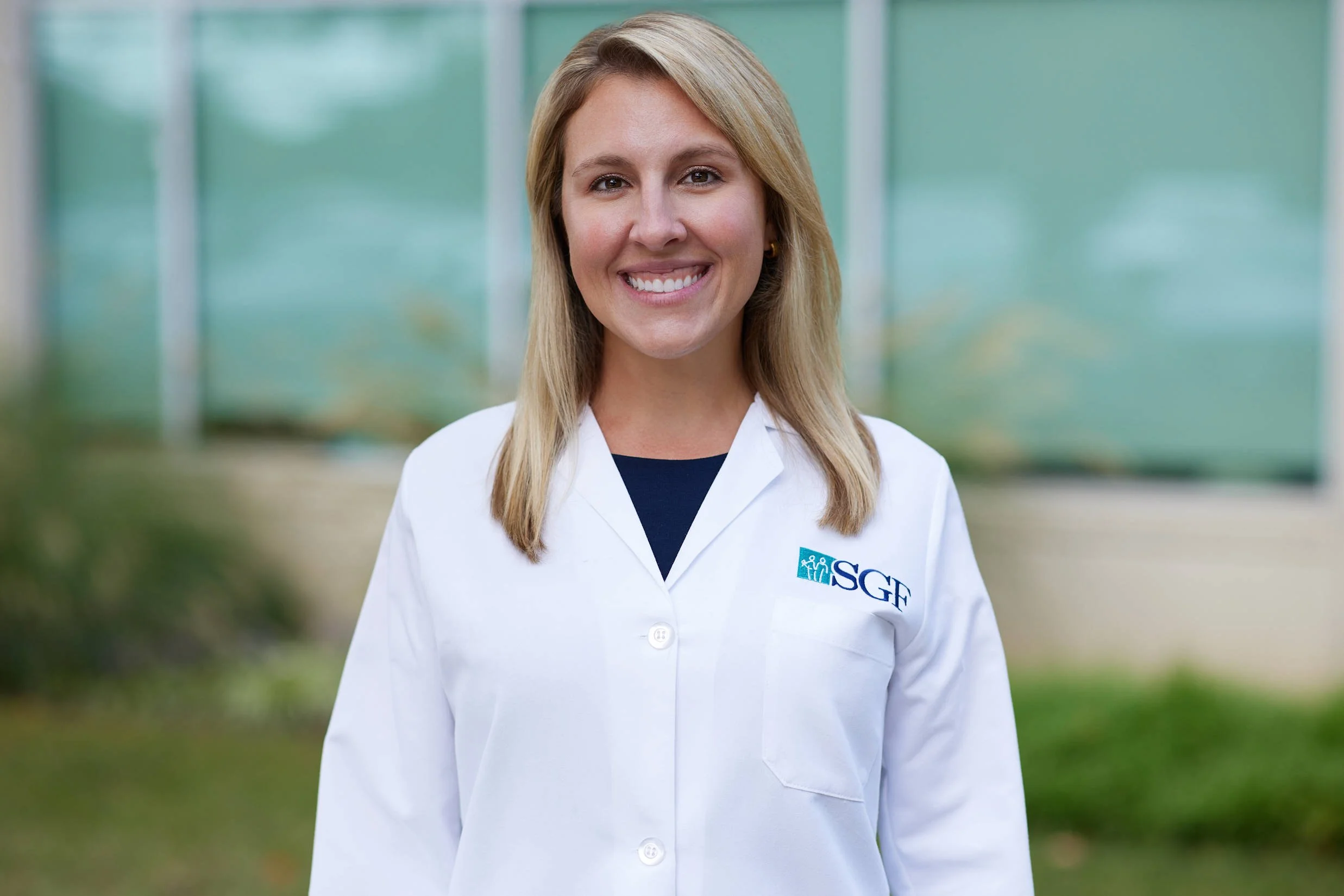 A woman in a white lab coat with "SGF" logo, standing outdoors, smiling.