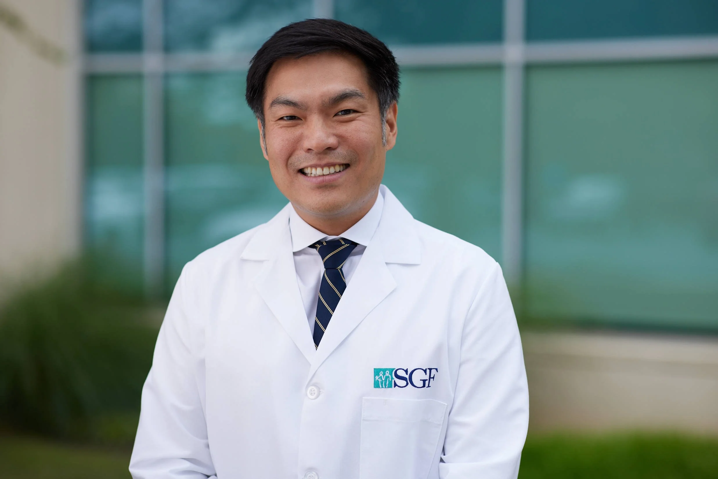 A person in a white lab coat with "SGF" logo standing outside a building, smiling.